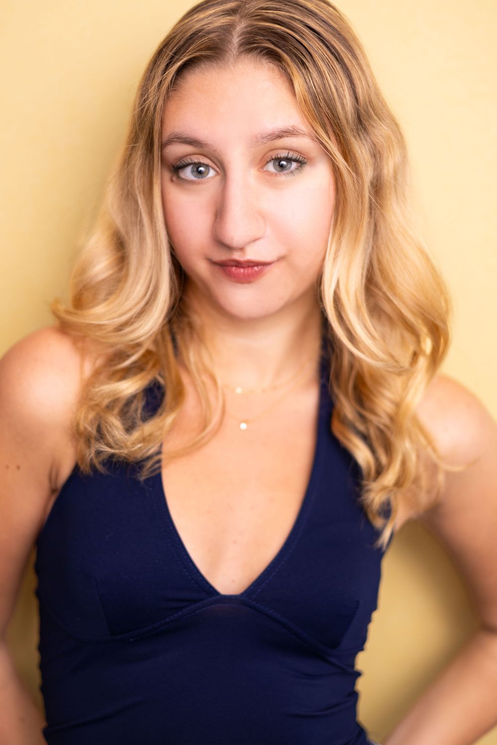 Young blonde woman with wavy hair in a navy halter top poses confidently against a yellow background.