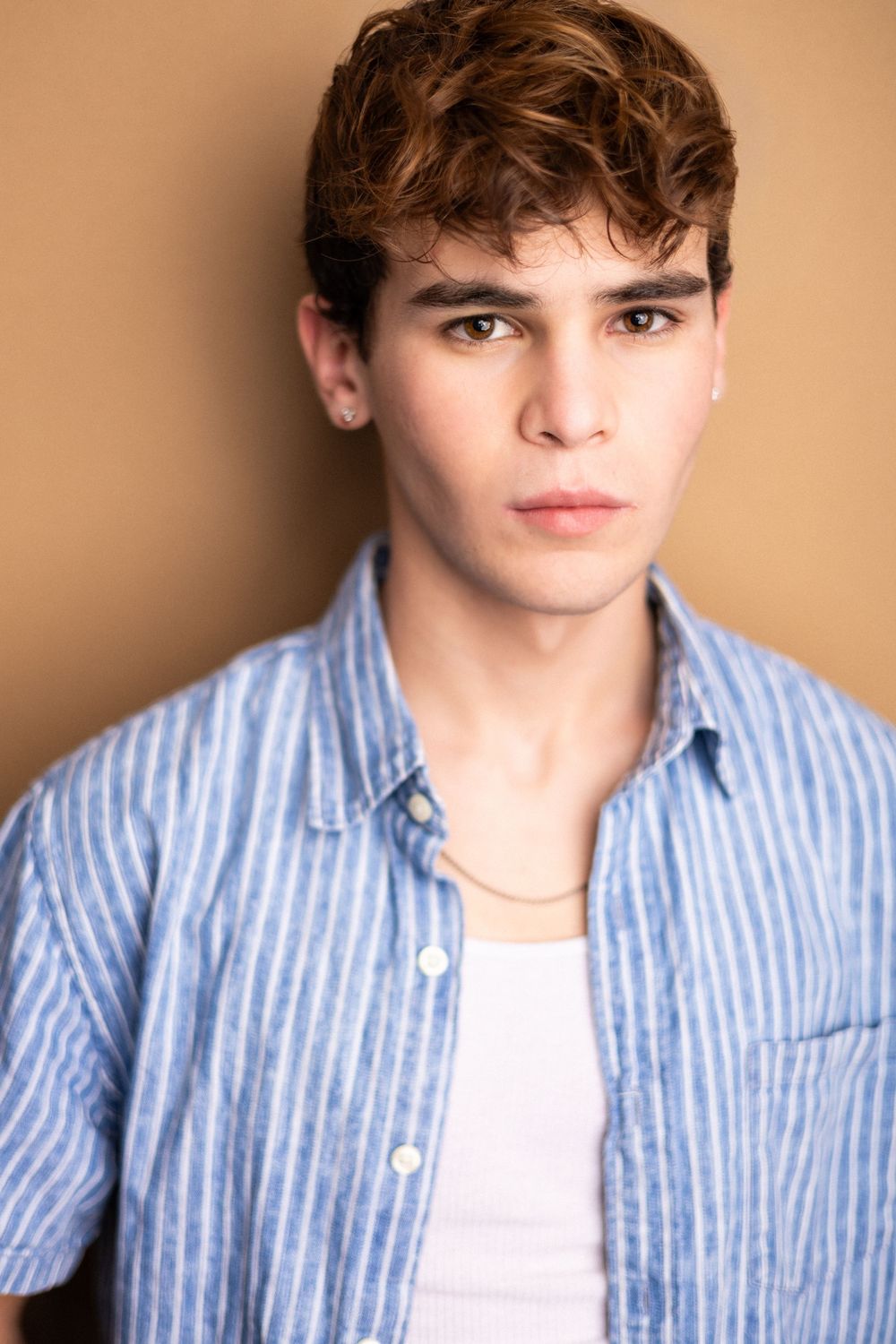 Young man with curly brown hair wearing a blue striped shirt and gold chain necklace against a beige background.