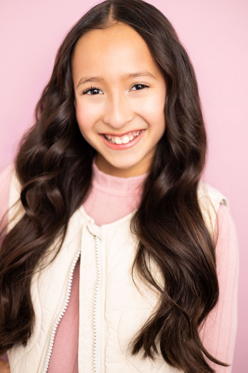 Happy young girl with long dark wavy hair wearing a white quilted vest over pink turtleneck on pink background.