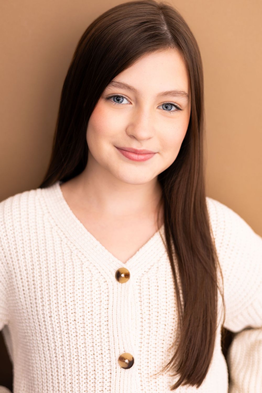 Young woman with long brown hair and blue eyes smiling, wearing a white knit sweater against a beige background.