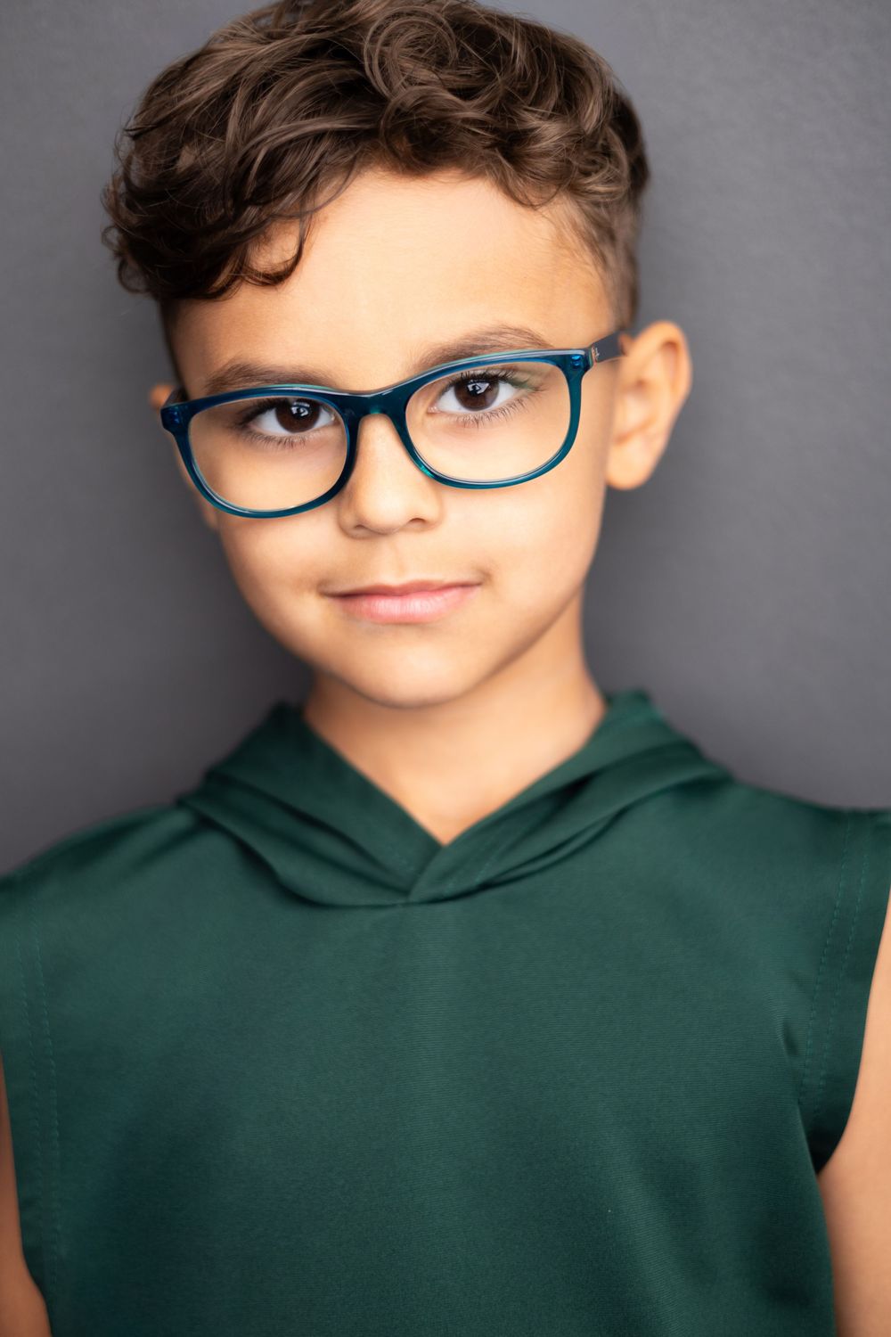 Young boy with curly hair wearing blue glasses and a green sleeveless hoodie, posing against a gray background.
