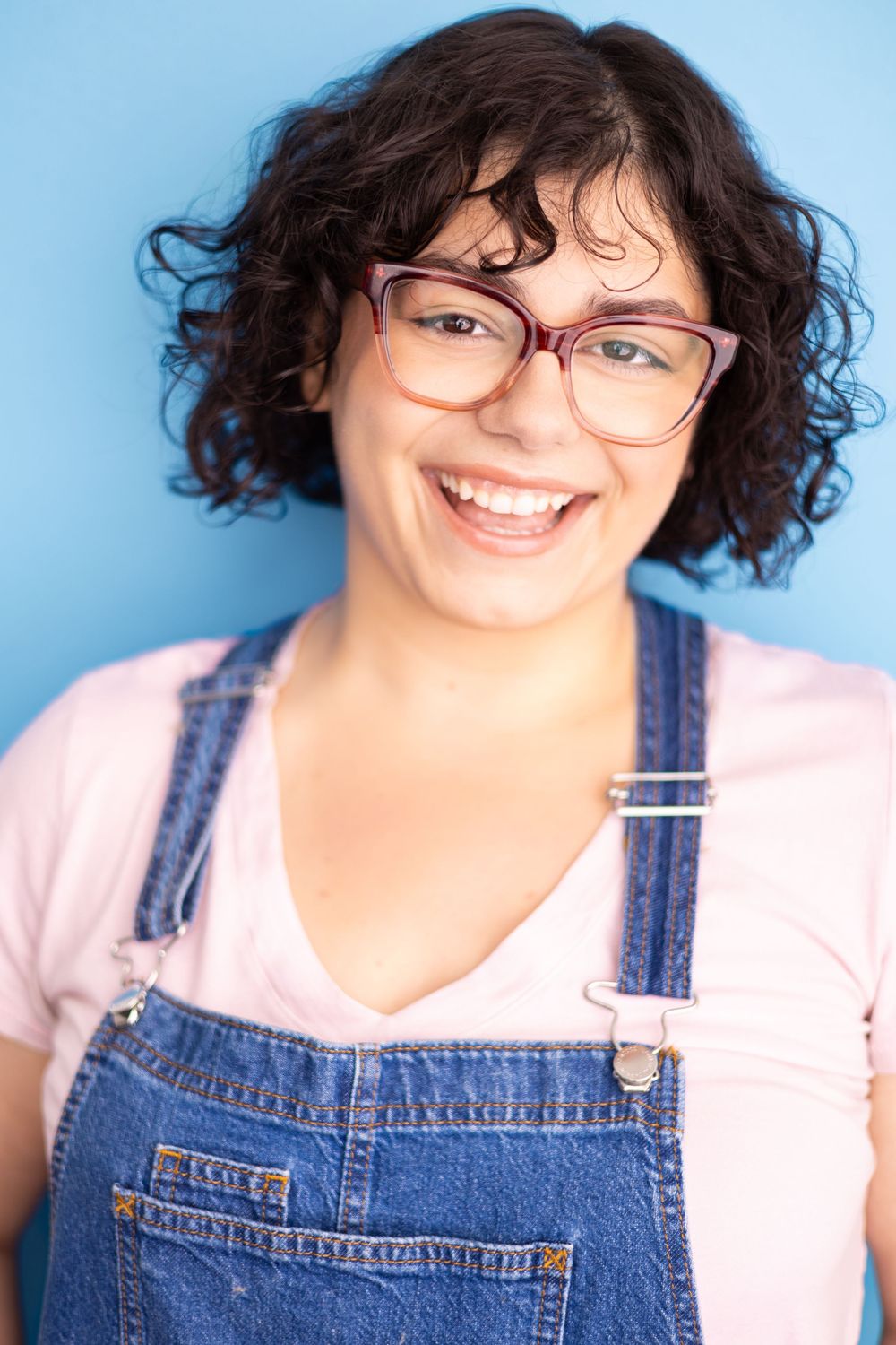 Smiling young woman with curly hair, glasses, and denim overalls poses against a bright blue background.
