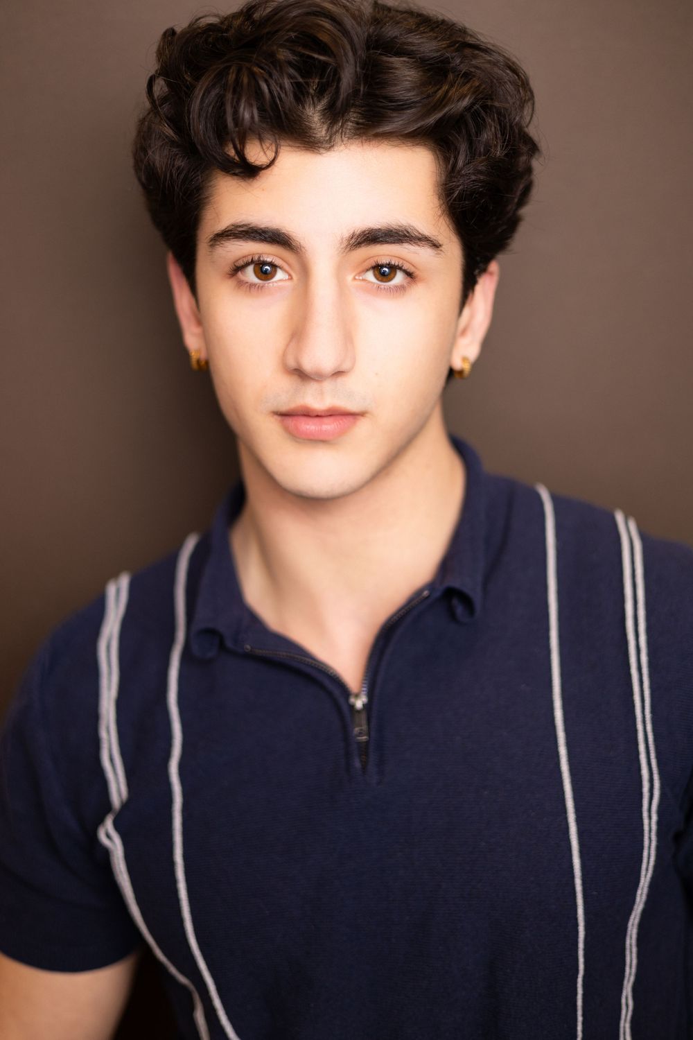 Serious young man with curly dark hair and gold stud earring wearing a navy striped zip polo shirt on a brown background.
