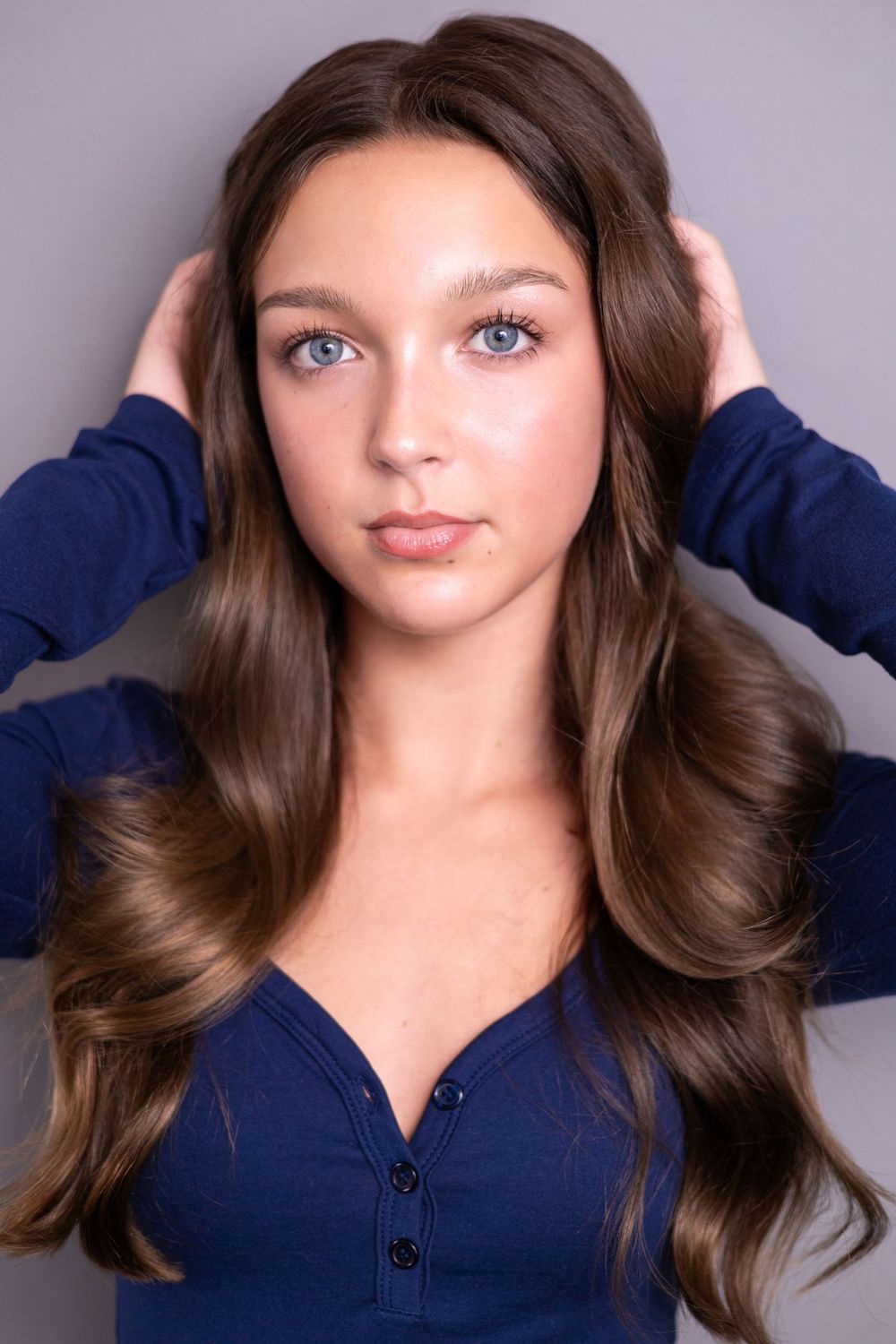A young woman with long wavy brown hair and blue eyes poses in a navy blue top against gray background.
