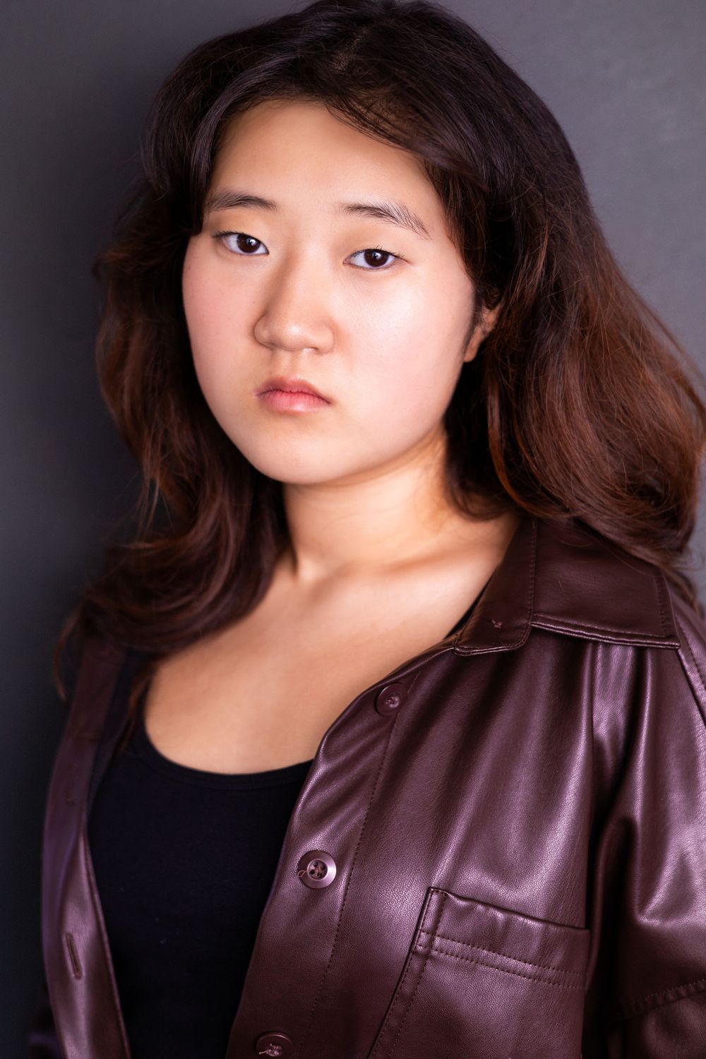 Young Asian woman with long brown hair poses seriously in a burgundy leather jacket against gray background.