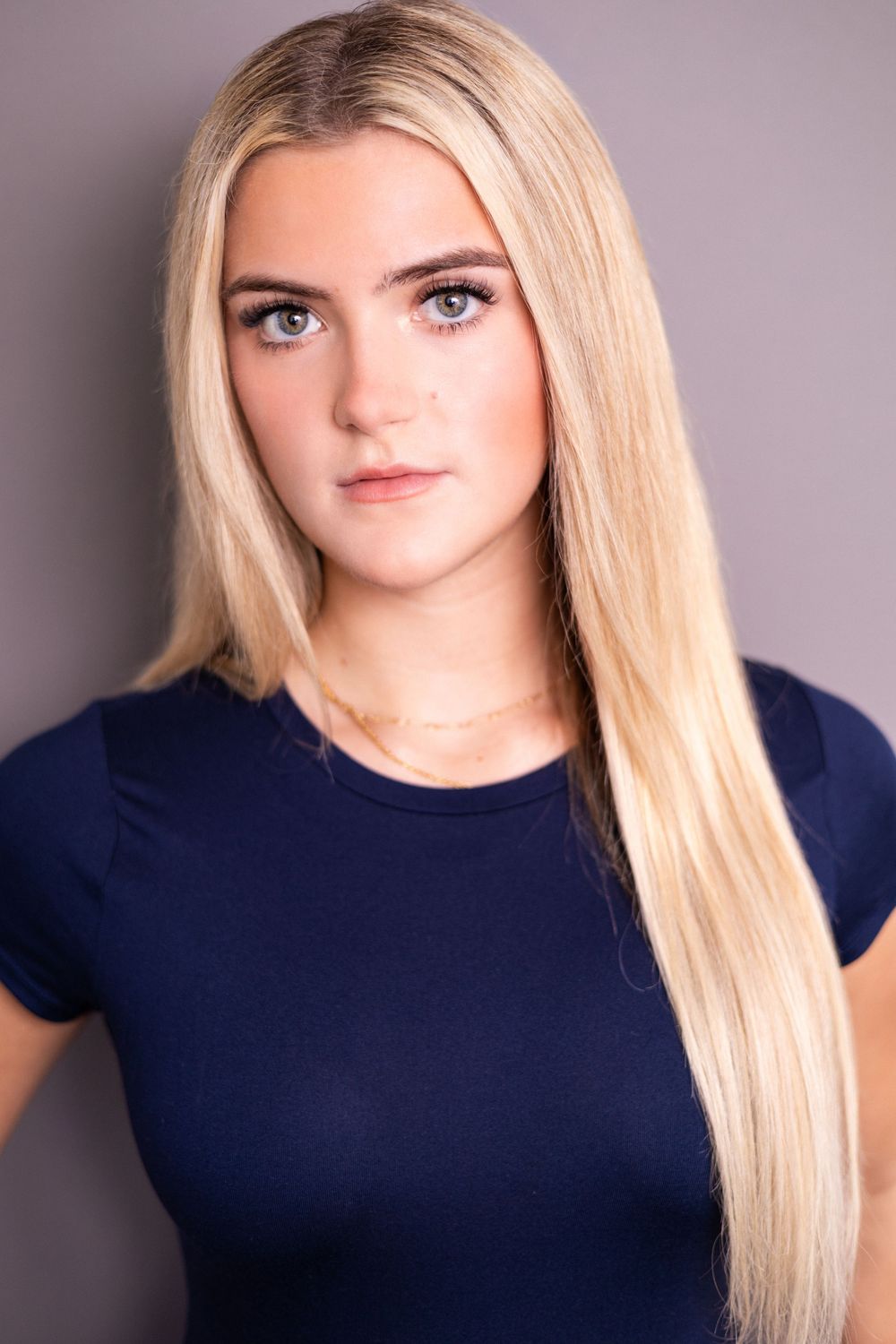 Young blonde woman with long straight hair wearing a navy blue top against a gray background.