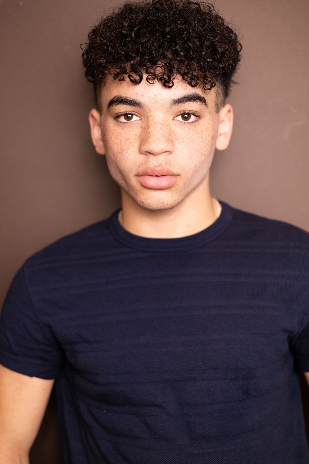 Young man with curly hair and freckles wearing a navy striped t-shirt poses against a brown background.