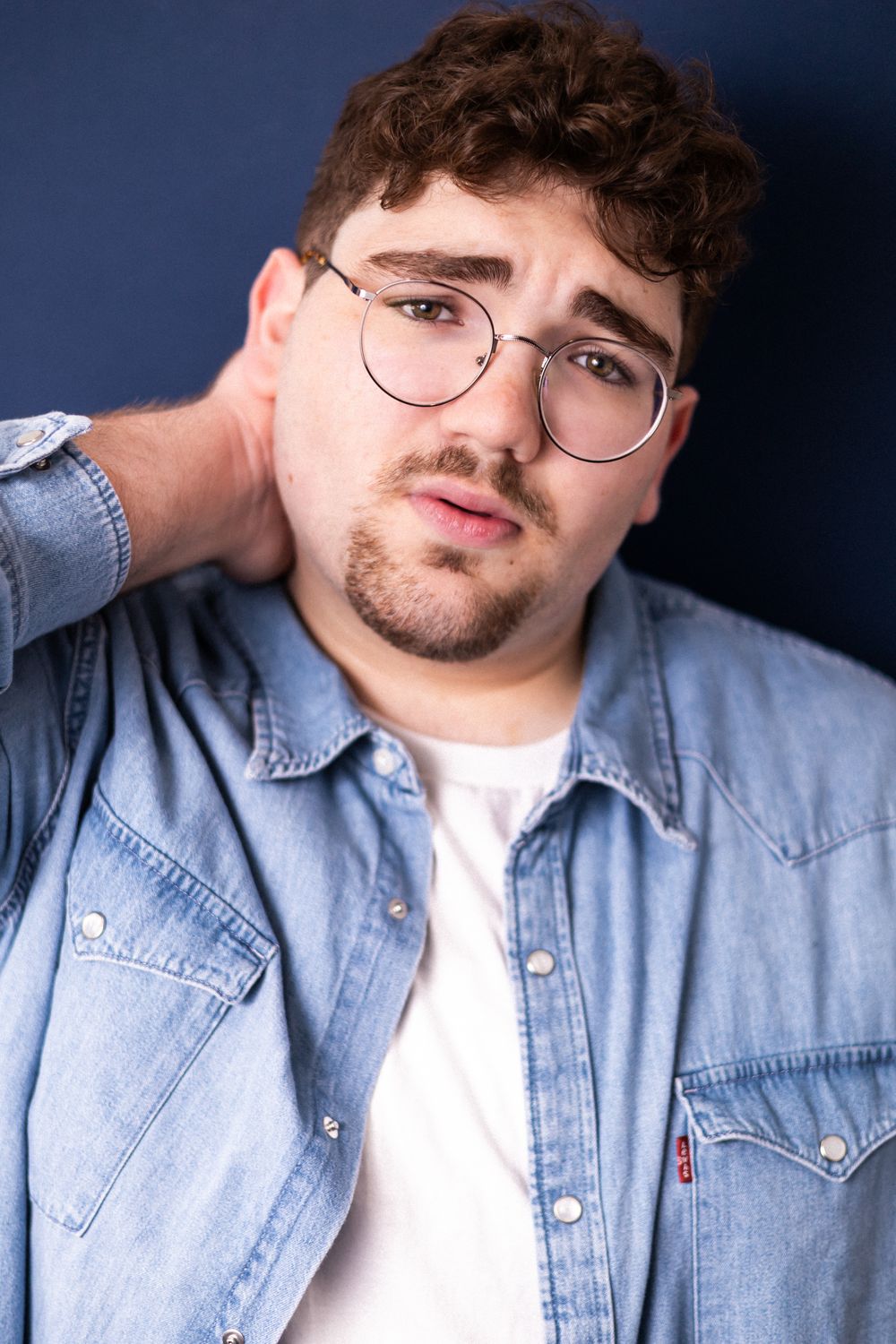 Young man with curly hair and glasses in a denim shirt touching his neck against a dark blue background.