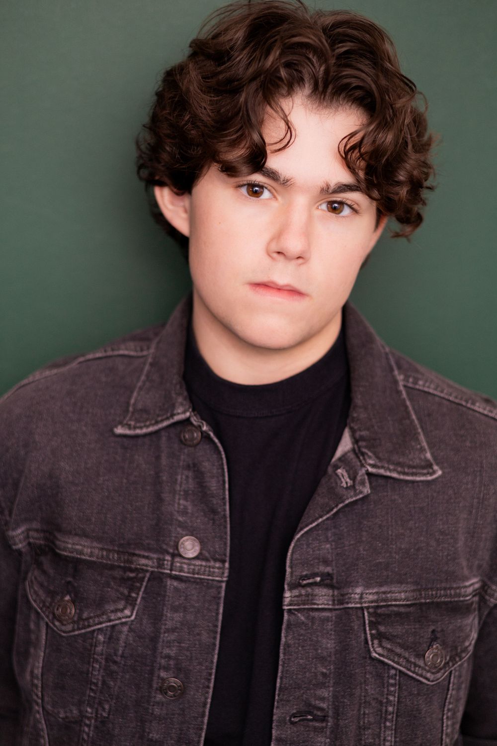 Young man with curly brown hair in a gray denim jacket poses seriously against a green background.