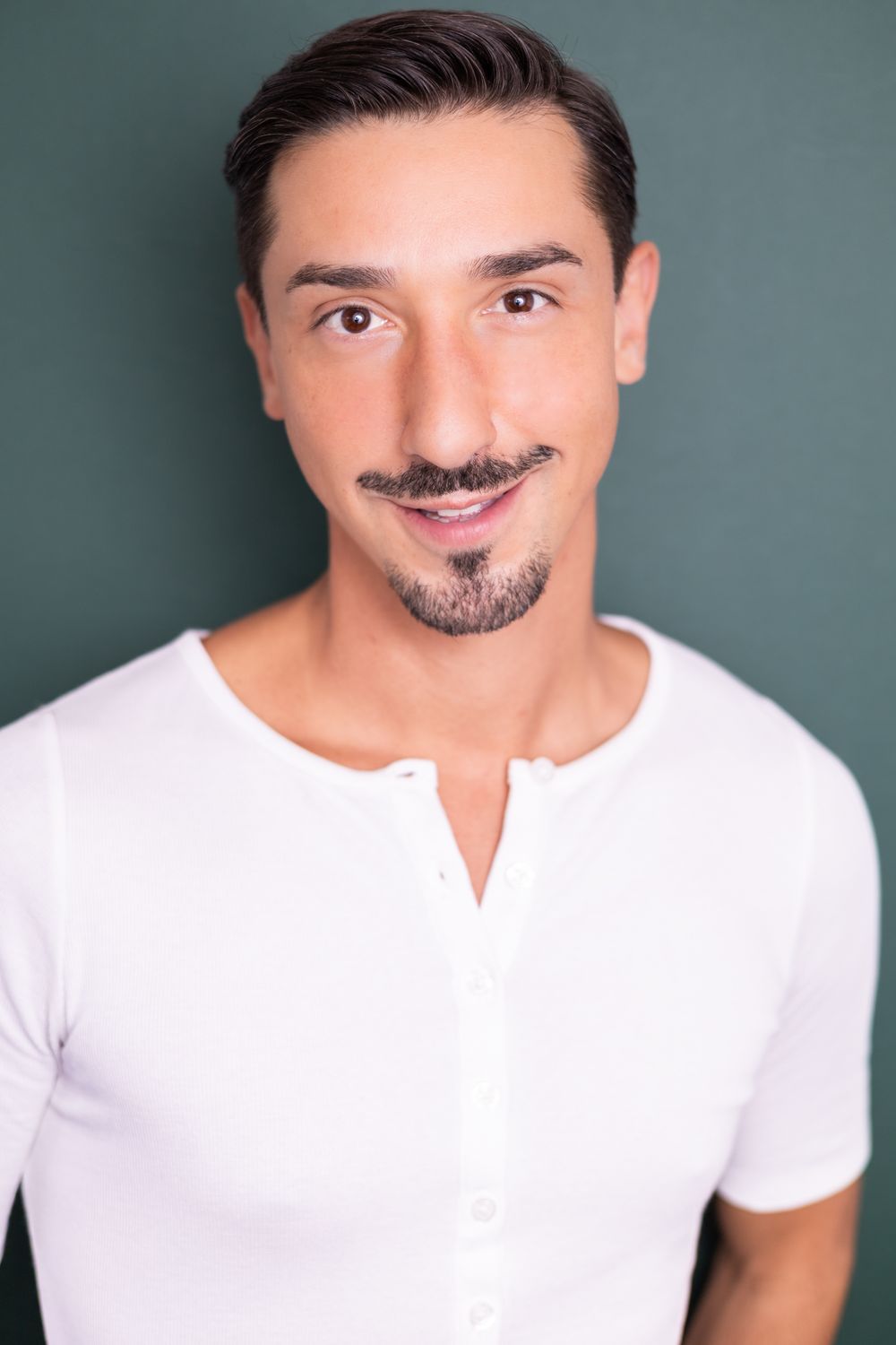 Young man with a goatee wearing a white henley shirt smiling against a green background.