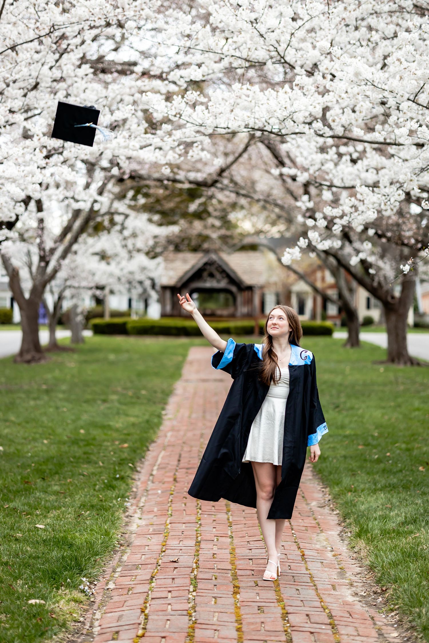 Graduate wearing a black cap and gown celebrates under blooming cherry blossom trees along a brick pathway in richmond, virginia RVA