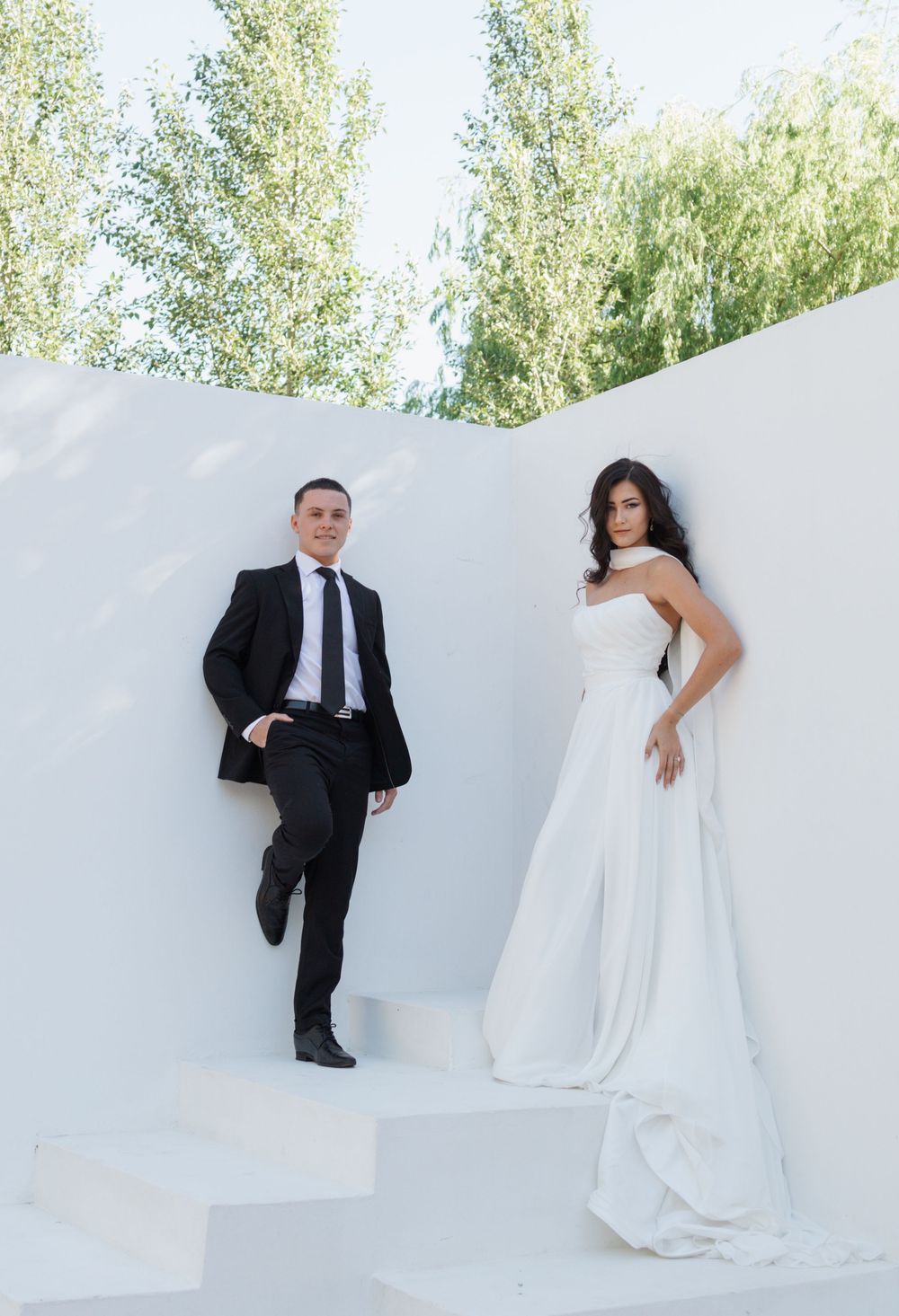 Bride in a flowing white gown and groom in a black suit pose on white steps against a white wall with tall green trees behind.