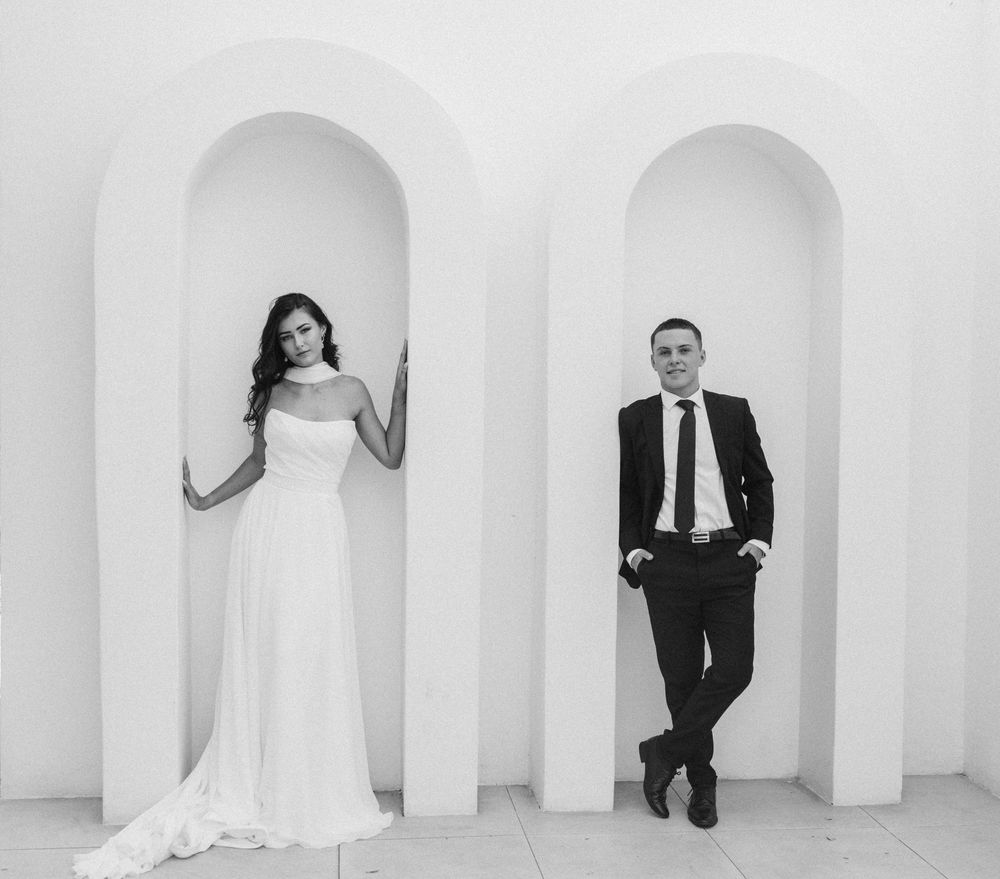 Black and white photo of a bride in a white gown and a groom in a suit posing in separate white arched alcoves.