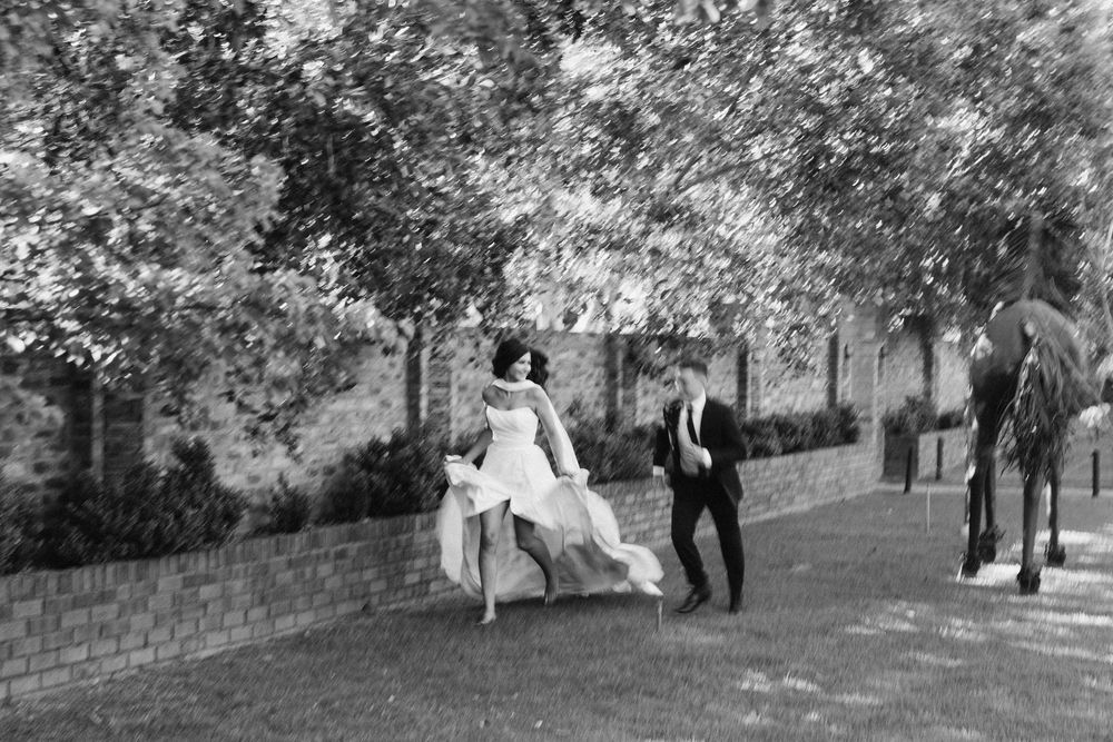 Black and white photo of a bride in a flowing gown running across a tree-shaded lawn toward a groom in a suit, with a horse nearby.