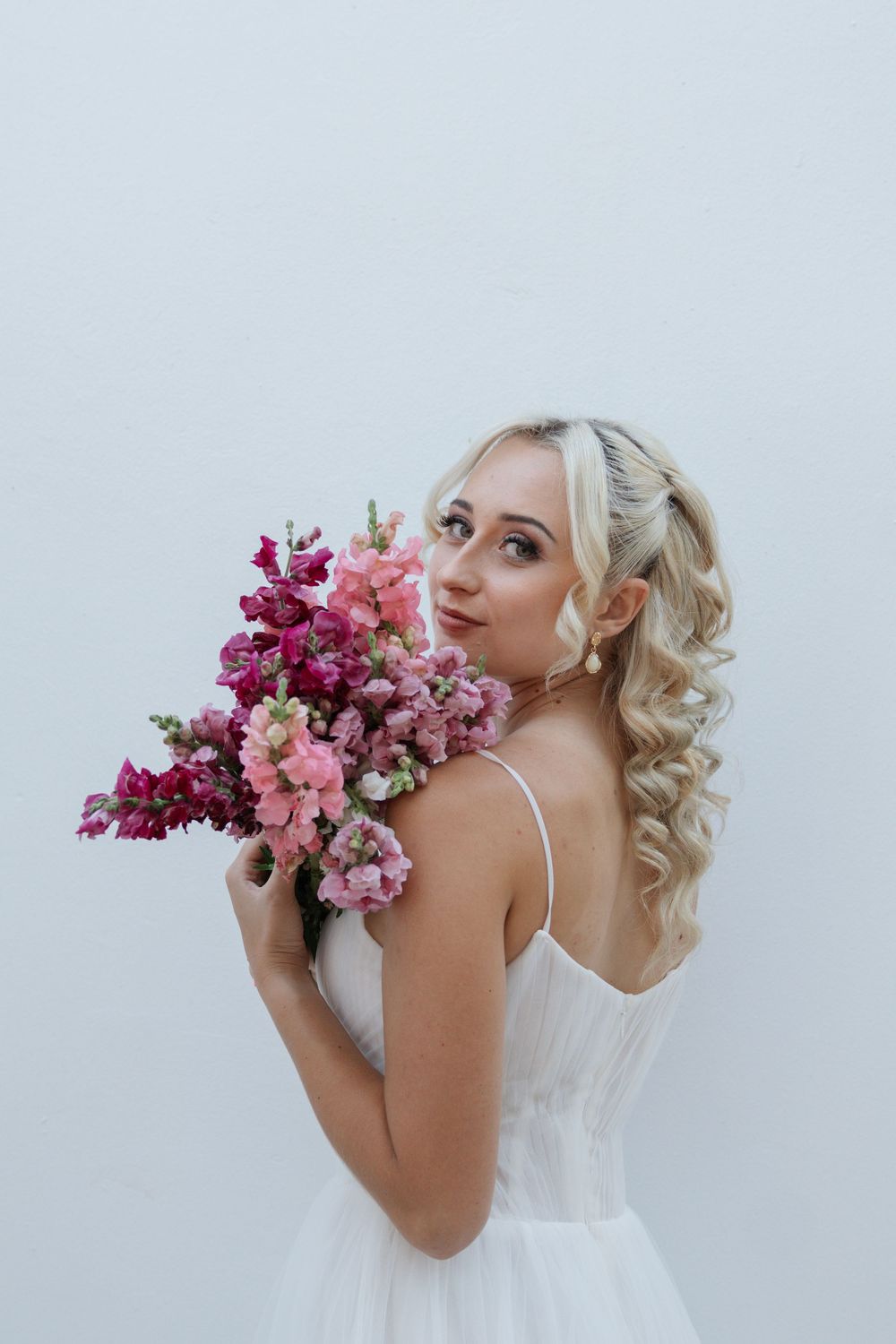 Bride in a white dress with blonde curled half-up hair glances over her shoulder holding a pink and magenta snapdragon bouquet.