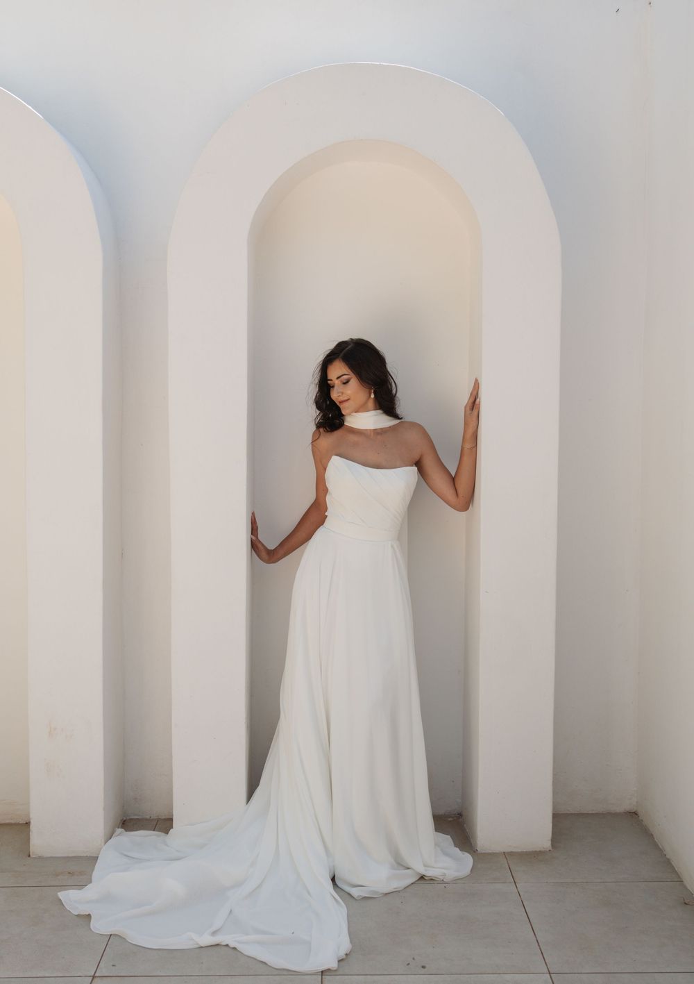 Bride in an elegant ivory strapless wedding dress with a high collar and long train, posing in a white arched alcove.