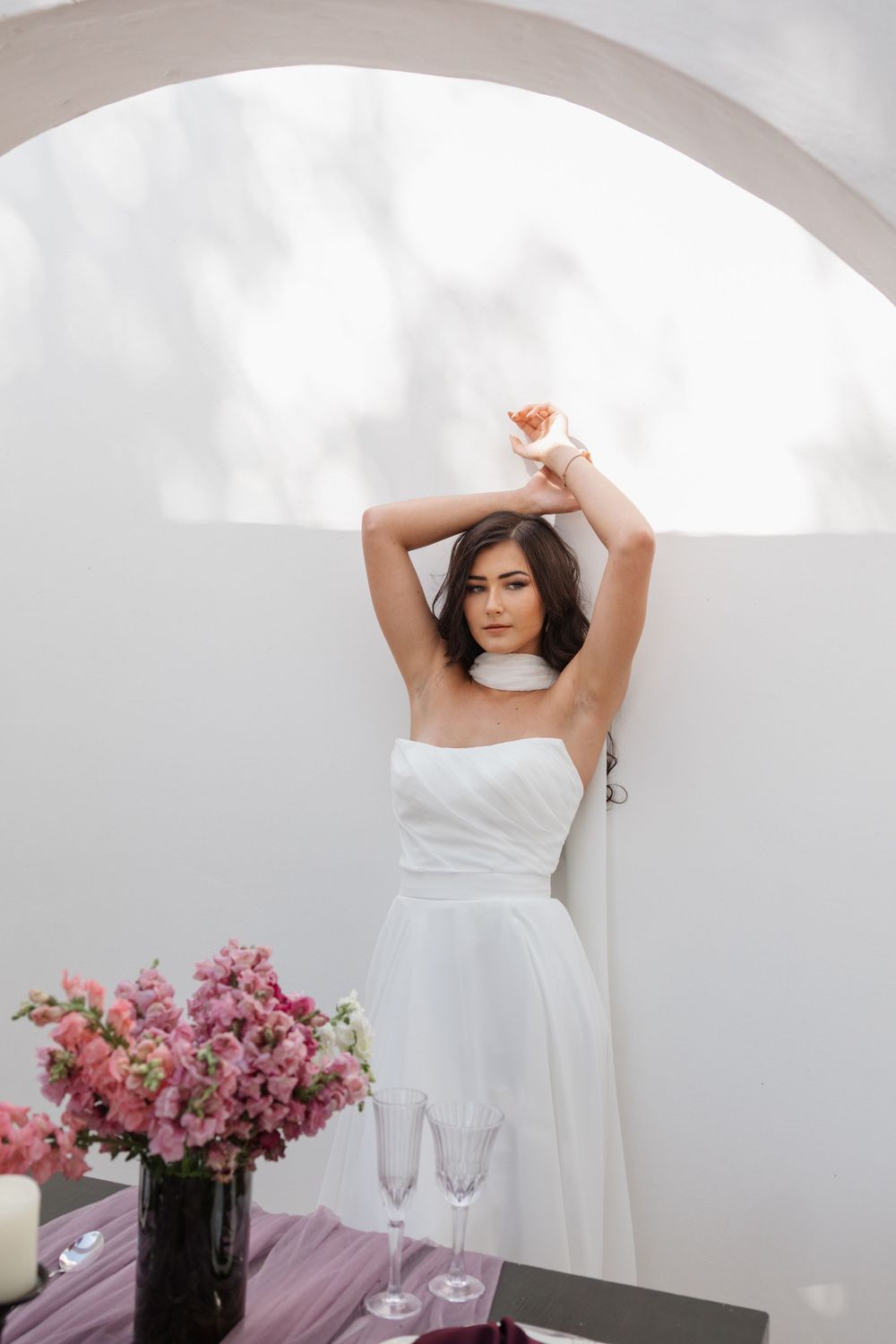 Woman in a white strapless bridal dress poses with arms raised beside a white arch, with pink flowers and champagne glasses on a table.