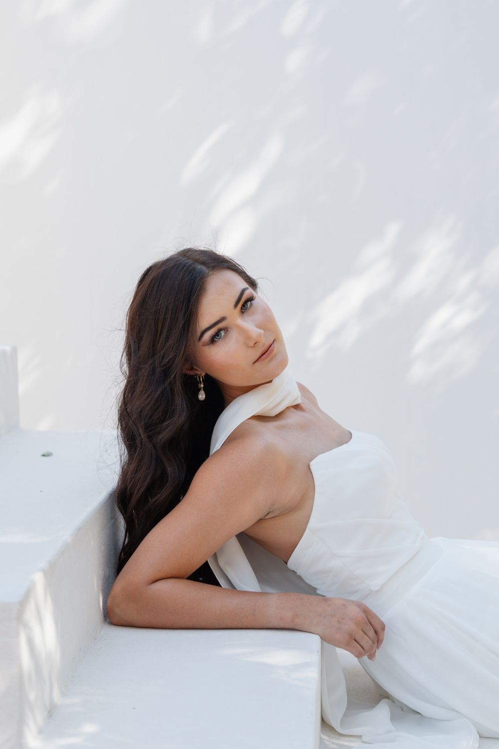 Woman with long dark wavy hair in an elegant white gown and drop earrings reclines on white steps in soft dappled sunlight.