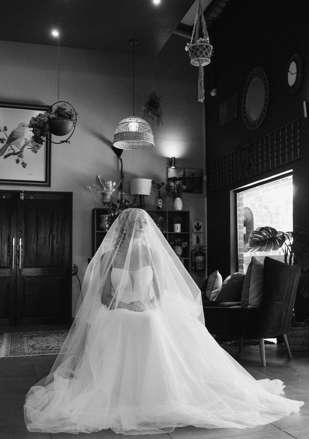 Bride in a flowing tulle gown and sheer veil seated in a cozy room with hanging lamps, plants and a leather armchair, in black and white.