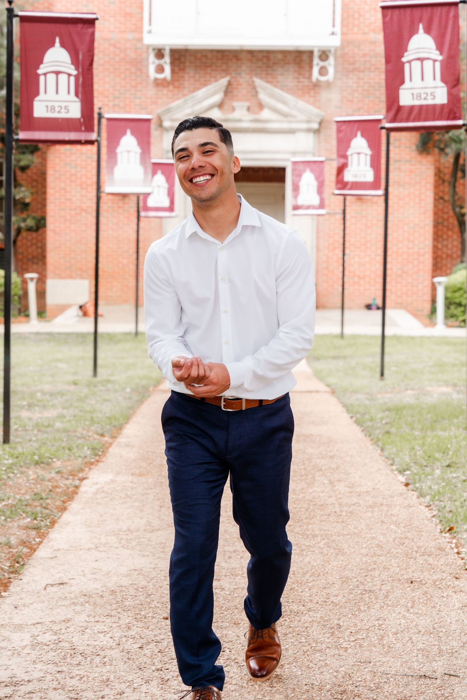 A person in a white dress shirt and navy pants walks along a brick path with red banners and university buildings in background.