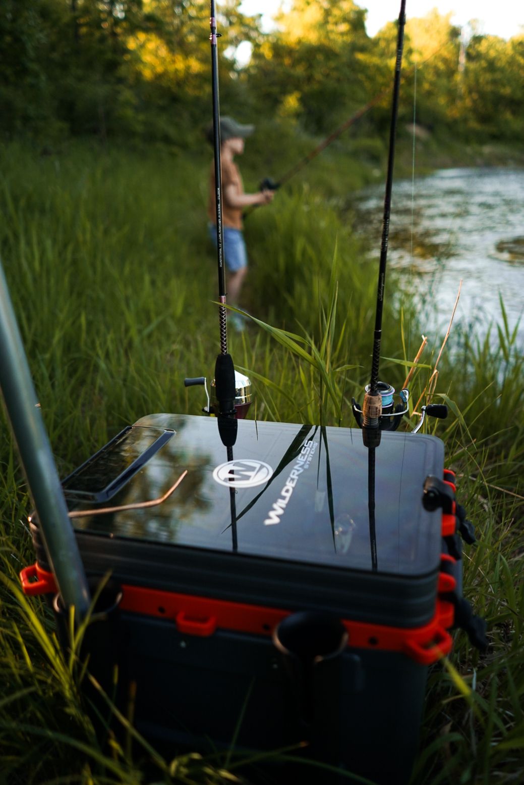 Fishing rods mounted on a tackle box by a riverbank during sunset.