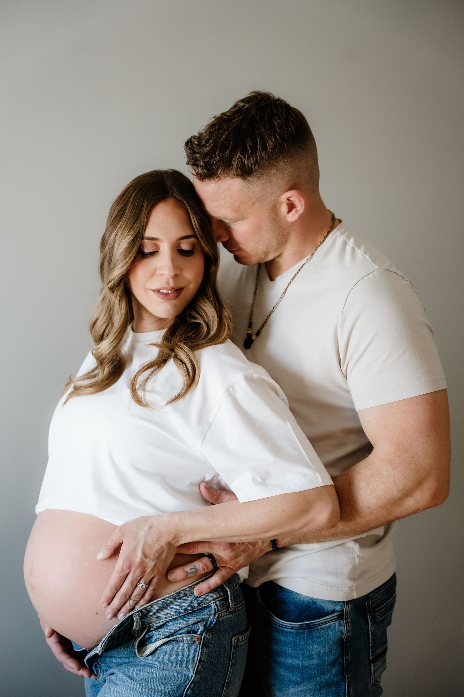 Intimate maternity photo of expectant couple in white shirts embracing against neutral wall. In home life style maternity session.