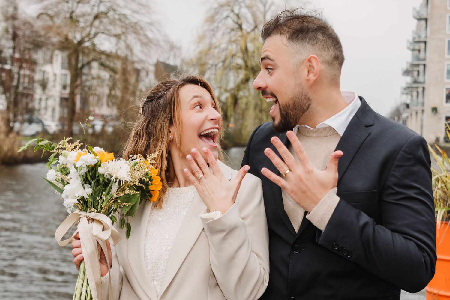 A joyful bridal couple shares an excited moment by a waterfront while holding a bouquet of white flowers after Stadhius wedding in Amsterdam