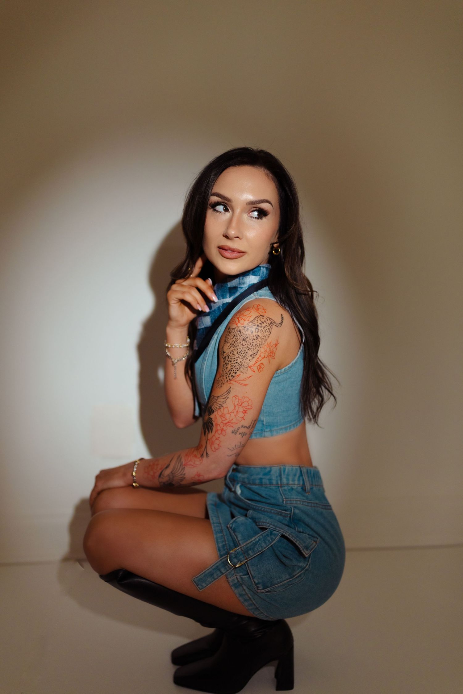 A person in a blue crop top and denim skirt poses crouched down against a neutral wall wearing black platform boots.