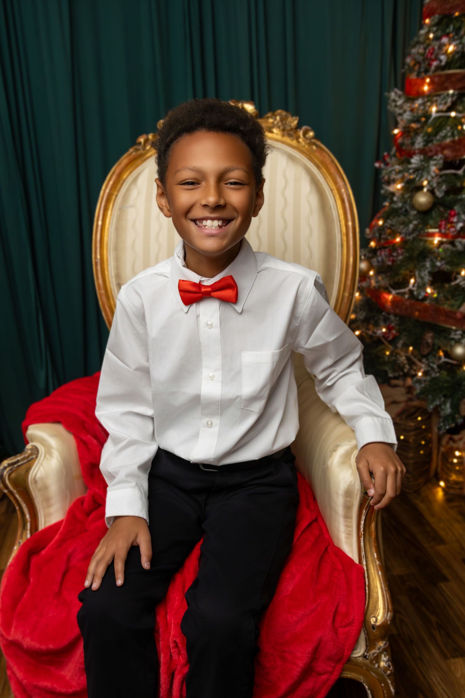A young man sitting on an ornate chair in a Christmas scene wearing a white collared shirt and a red bowtie
