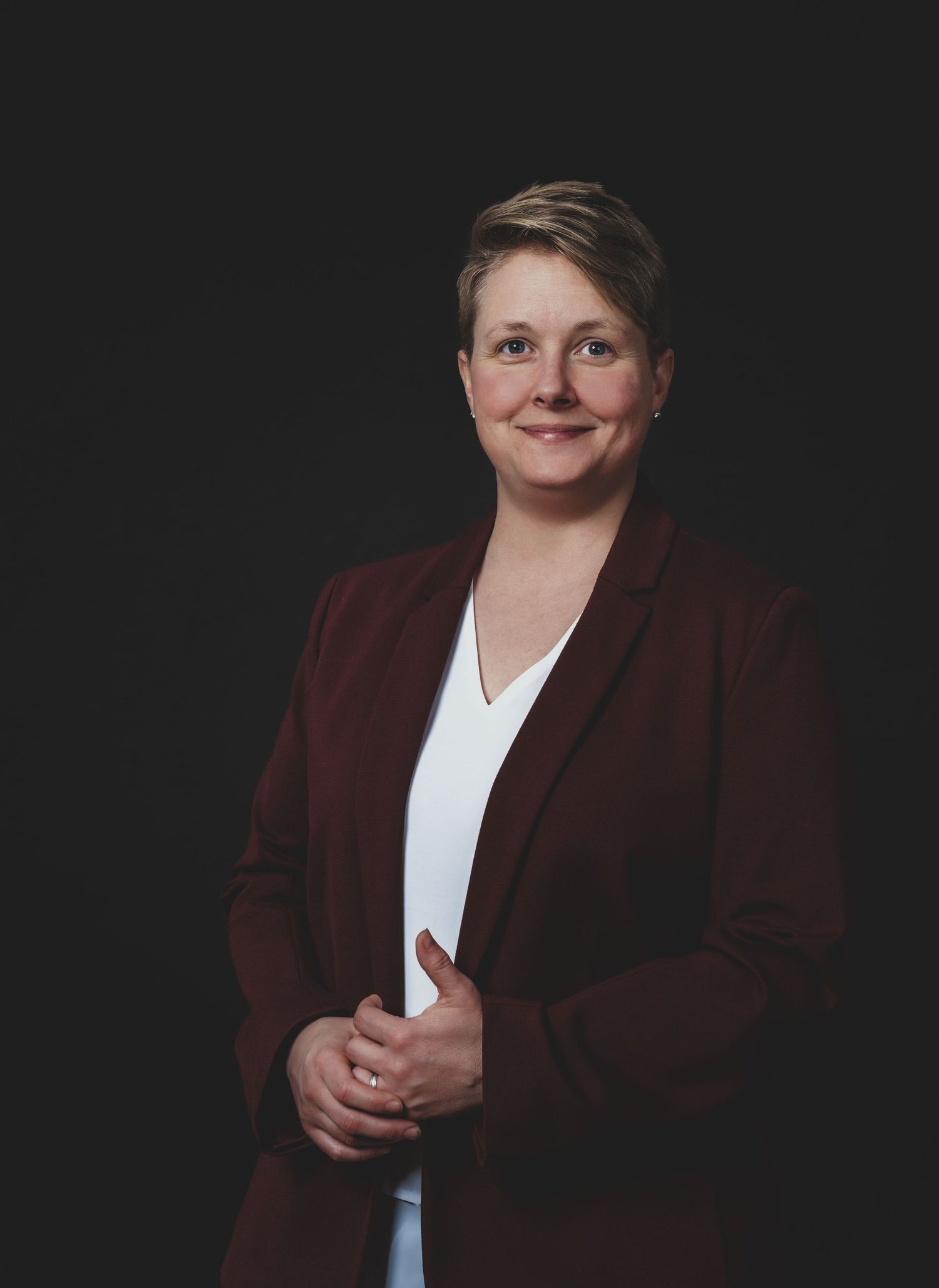 A professional portrait of a person in a burgundy blazer and white top against a dark background, smiling warmly.