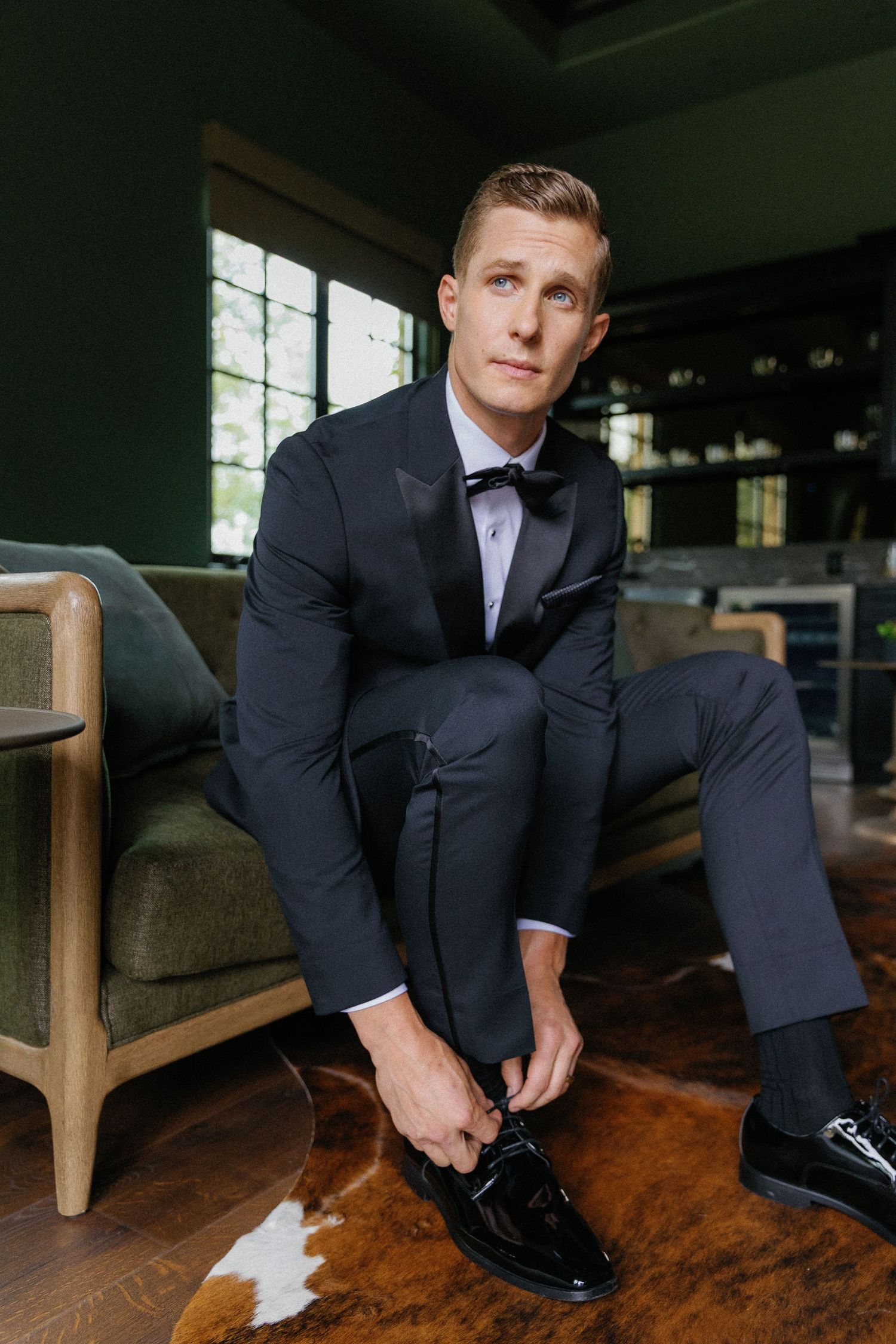 A groom in a classic black tuxedo and bowtie sits on a brown leather chair while tying his formal dress shoes.