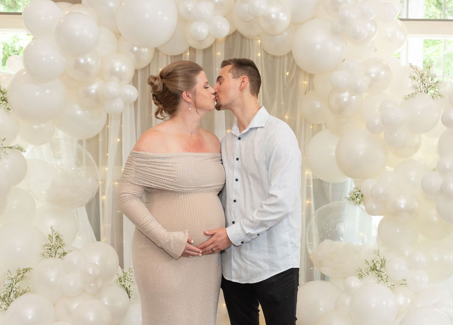 A couple poses in front of an elegant white balloon and floral wall display.