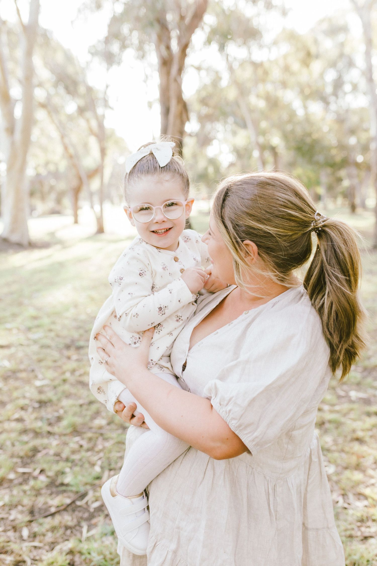 Woman in white dress holding smiling baby outdoors in natural sunlit setting.