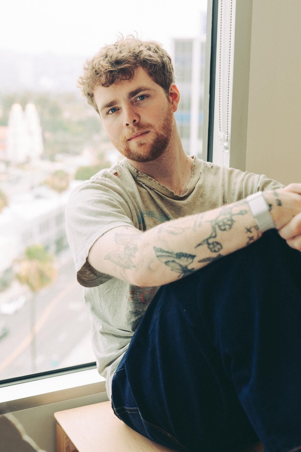 Someone poses alongside a large window in different casual seated positions wearing a light colored shirt.