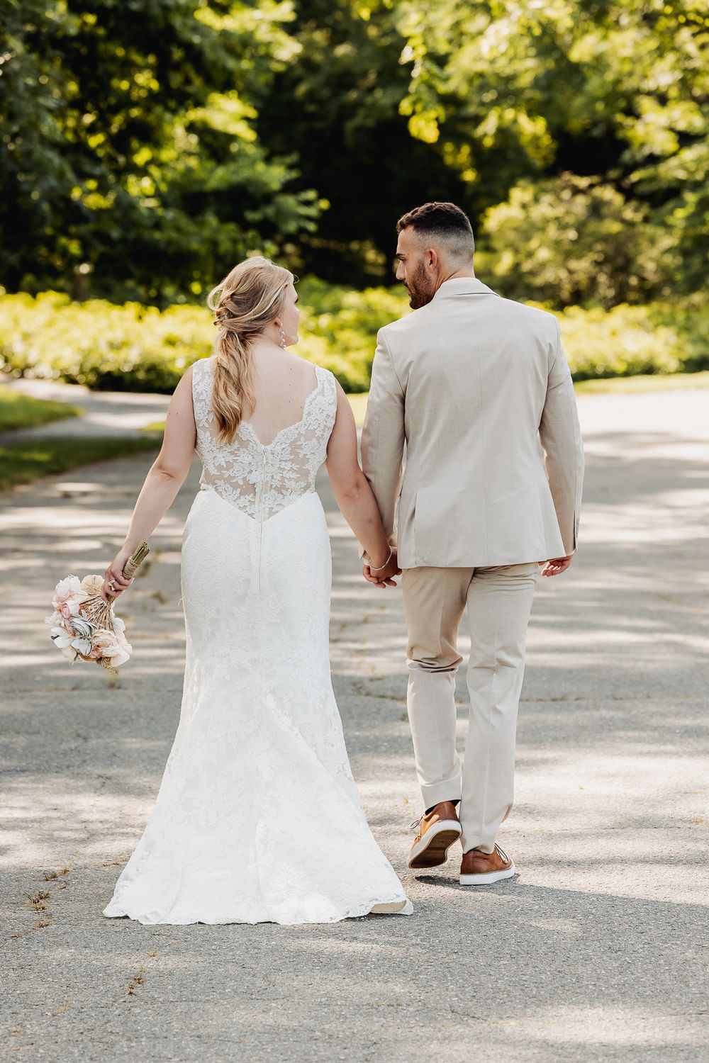 A couple in wedding attire walks away on a sunlit path holding hands during an outdoor summer ceremony.