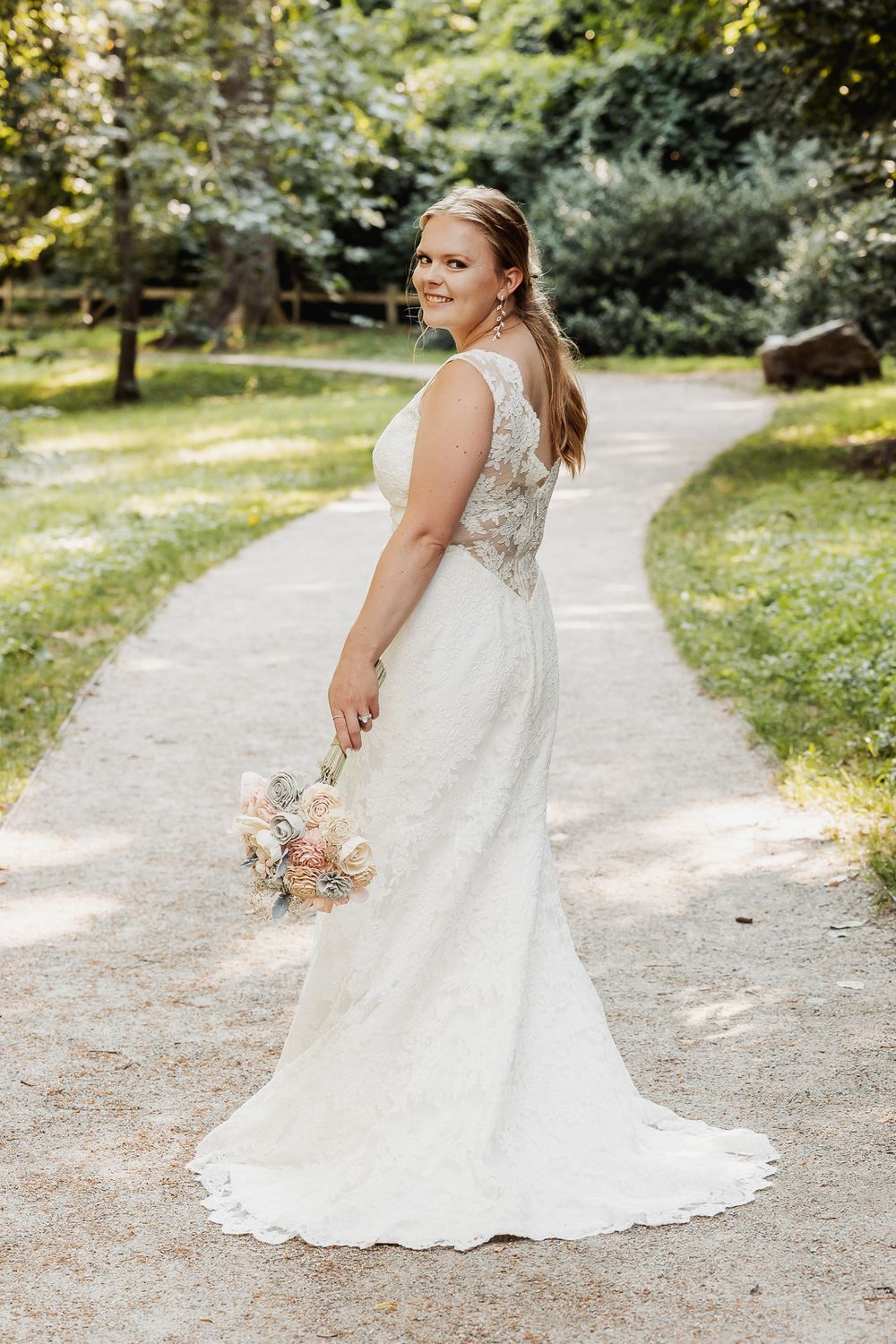 Elegant white lace wedding gown with detailed back shown on garden path surrounded by greenery.