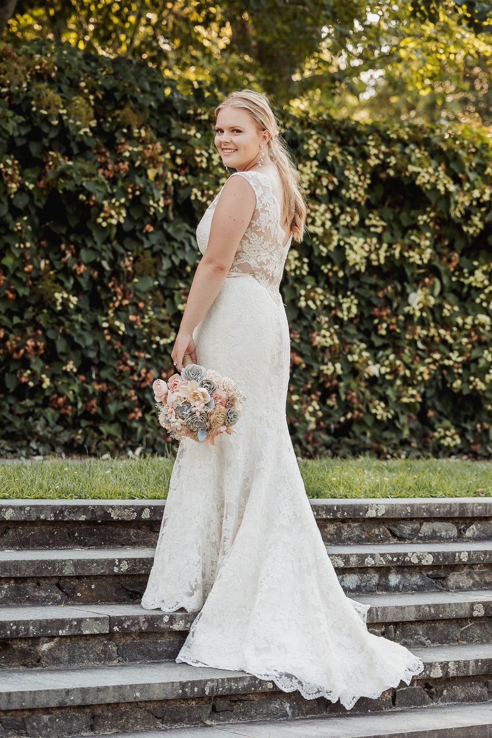 A bride in a white lace dress poses on garden stone steps while holding a pastel-colored floral bouquet.