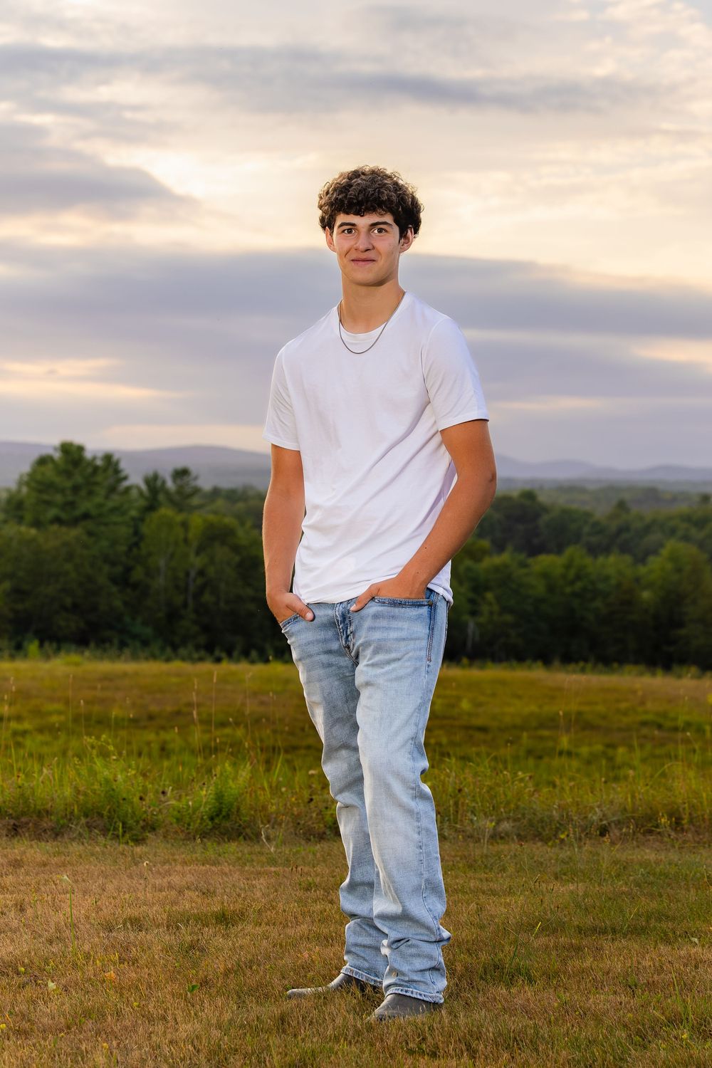 Person in casual white t-shirt and jeans poses outdoors in a grassy field at sunset with mountains in the background.