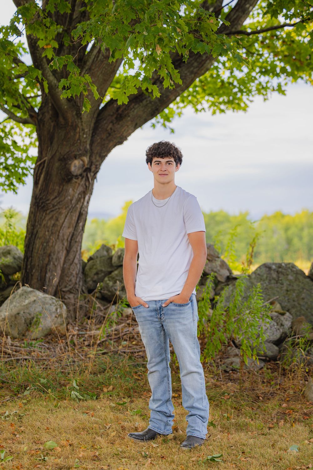 A person wearing jeans and white t-shirt poses near trees and stones in an outdoor nature setting during golden hour.