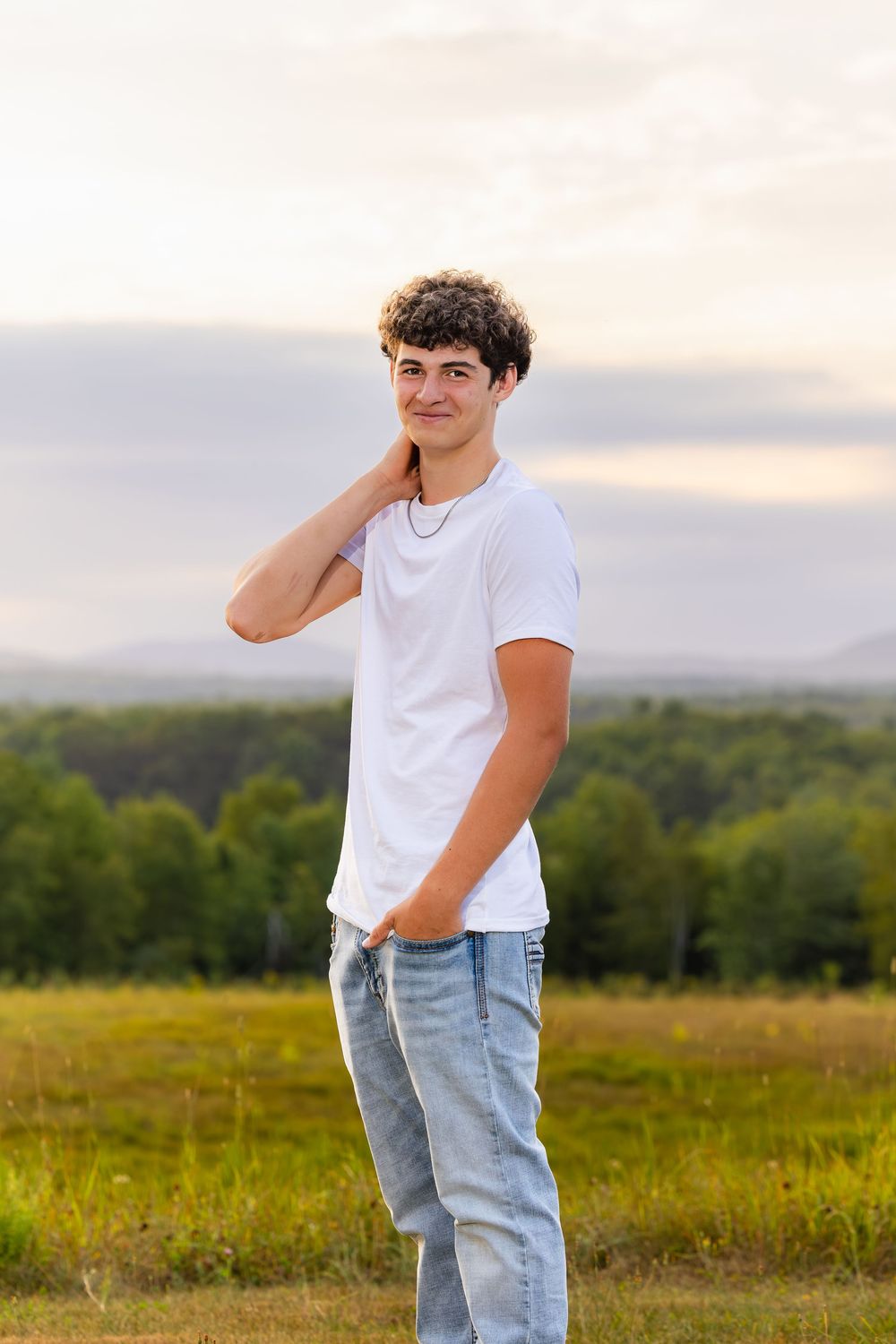 Someone in a white t-shirt and light blue jeans poses casually in a scenic meadow with rolling hills during sunset.