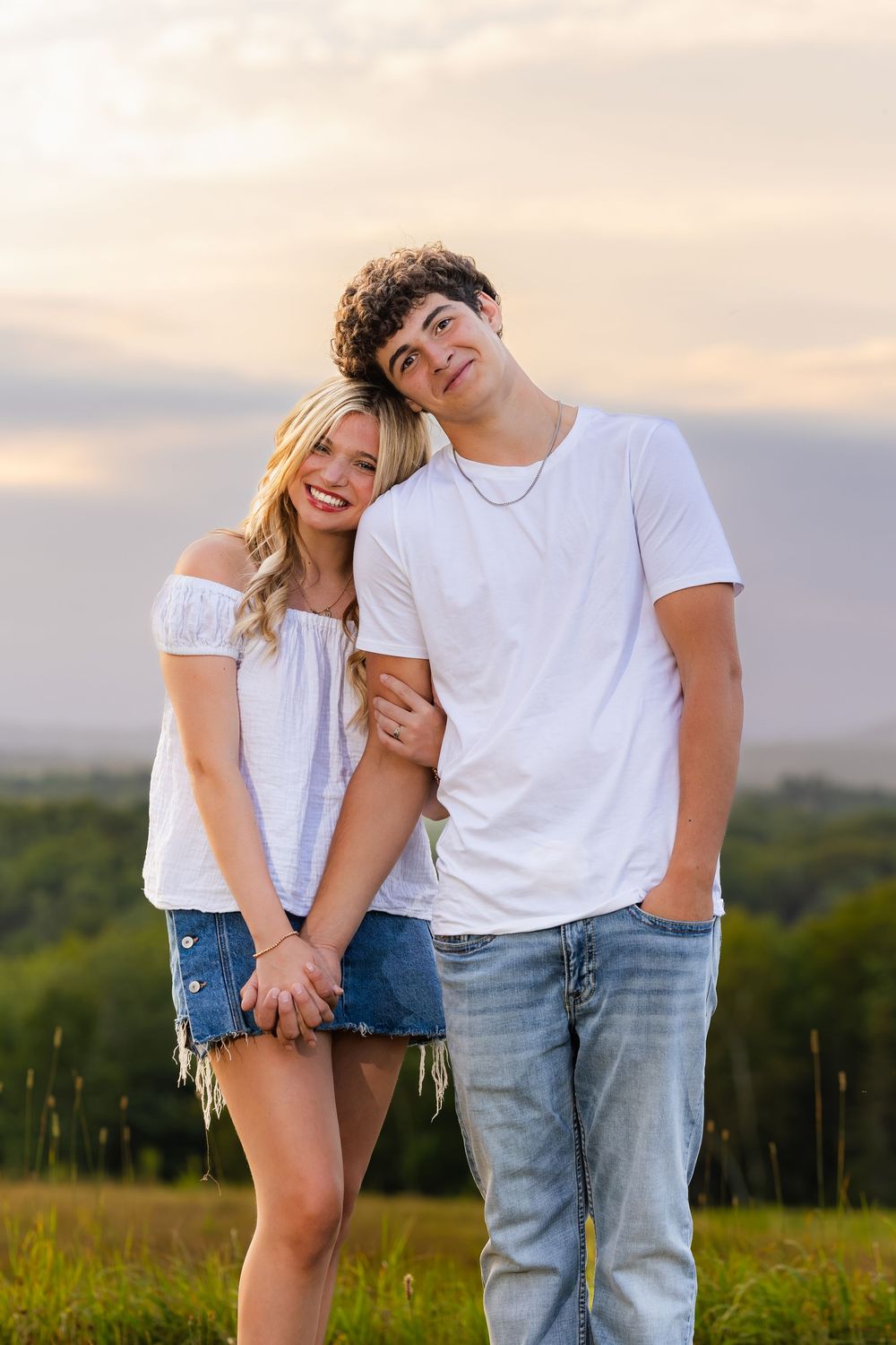Young couple in white tops posing together in a grassy field at sunset.