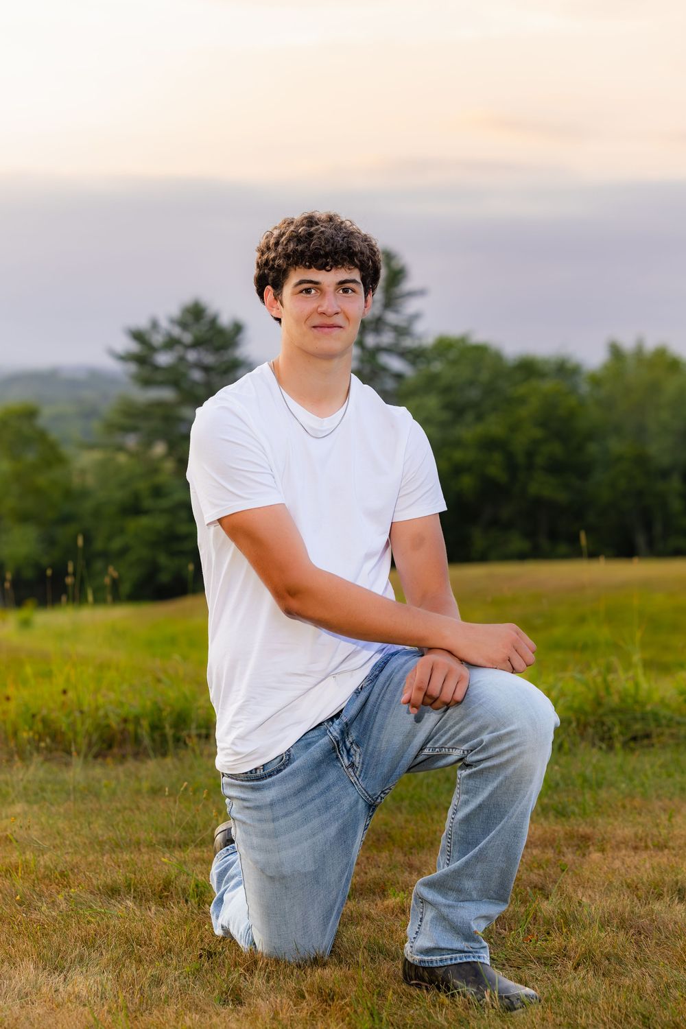 A young person in a white t-shirt and jeans sits casually in a grassy field at sunset.