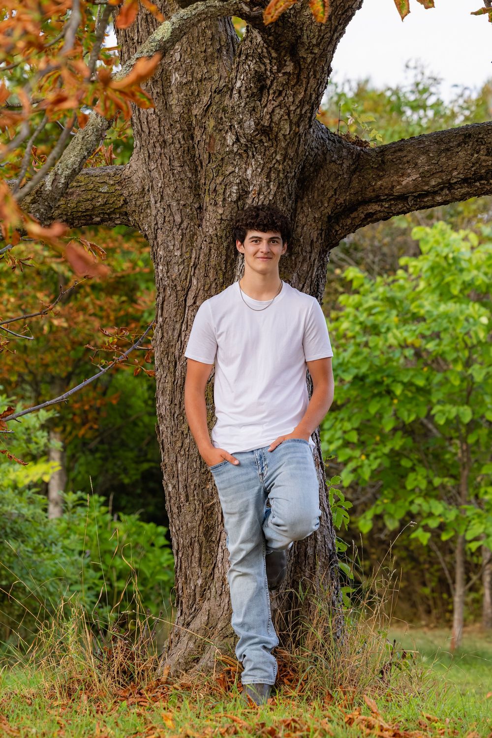 A person in a white t-shirt and jeans sits casually against the trunk of a large tree during autumn.