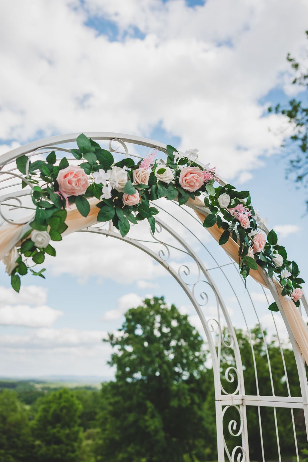 White metal wedding arch adorned with pink roses and green ivy vines against a bright blue cloudy sky.