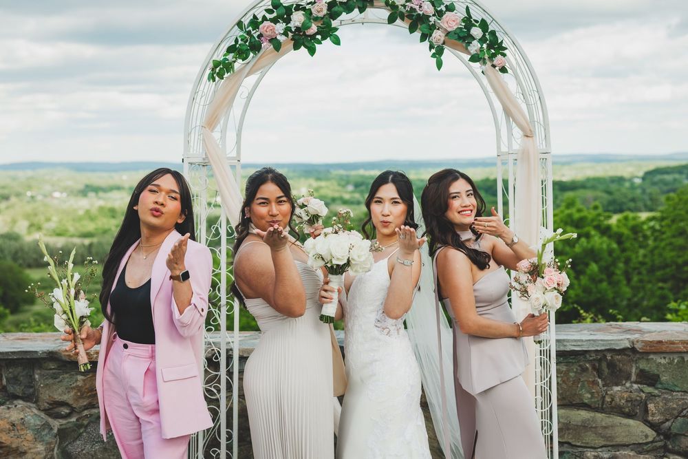 A group of bridesmaids pose together under a floral arch at an outdoor wedding venue with scenic rolling hills in background.