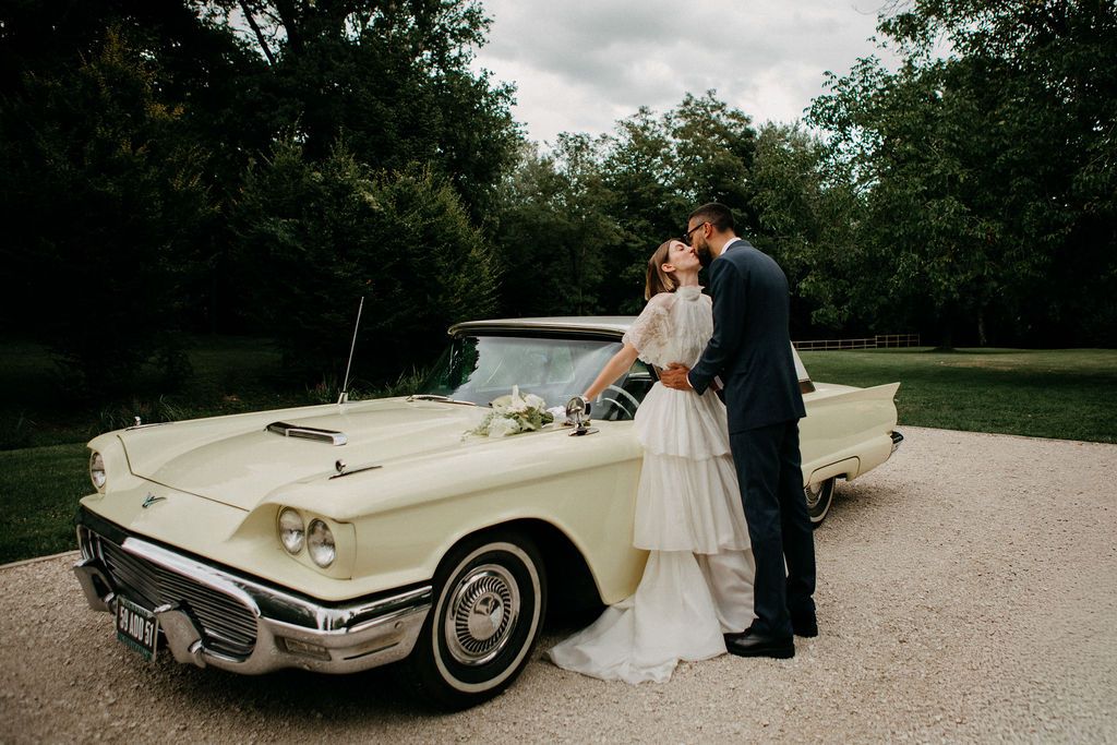 Bride and groom share a romantic kiss in front of a vintage cream Ford Thunderbird convertible.