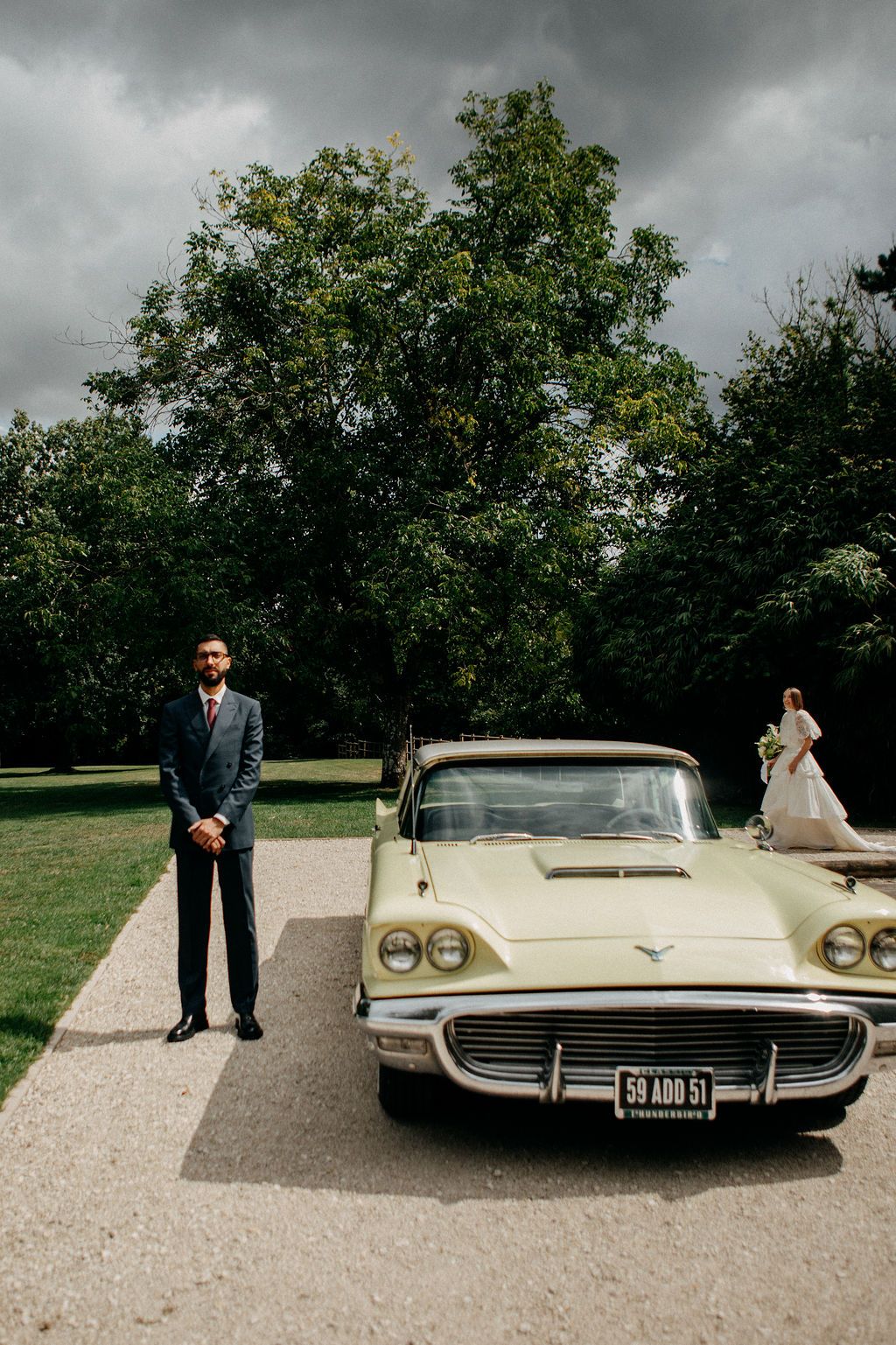 Groom in grey suit standing beside vintage yellow Ford Thunderbird with bride in background.