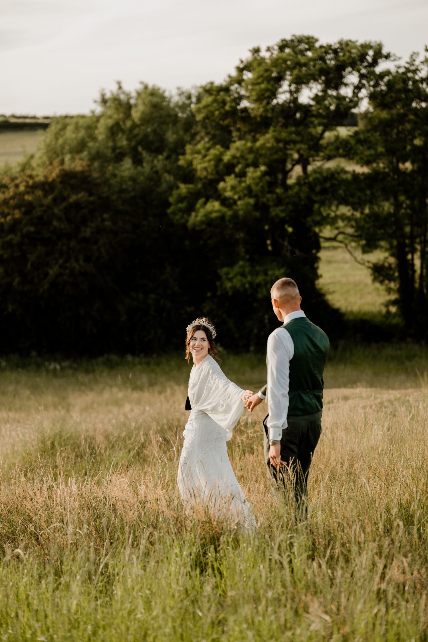 Newlyweds walking through tall grass in a sunny field during sunset.
