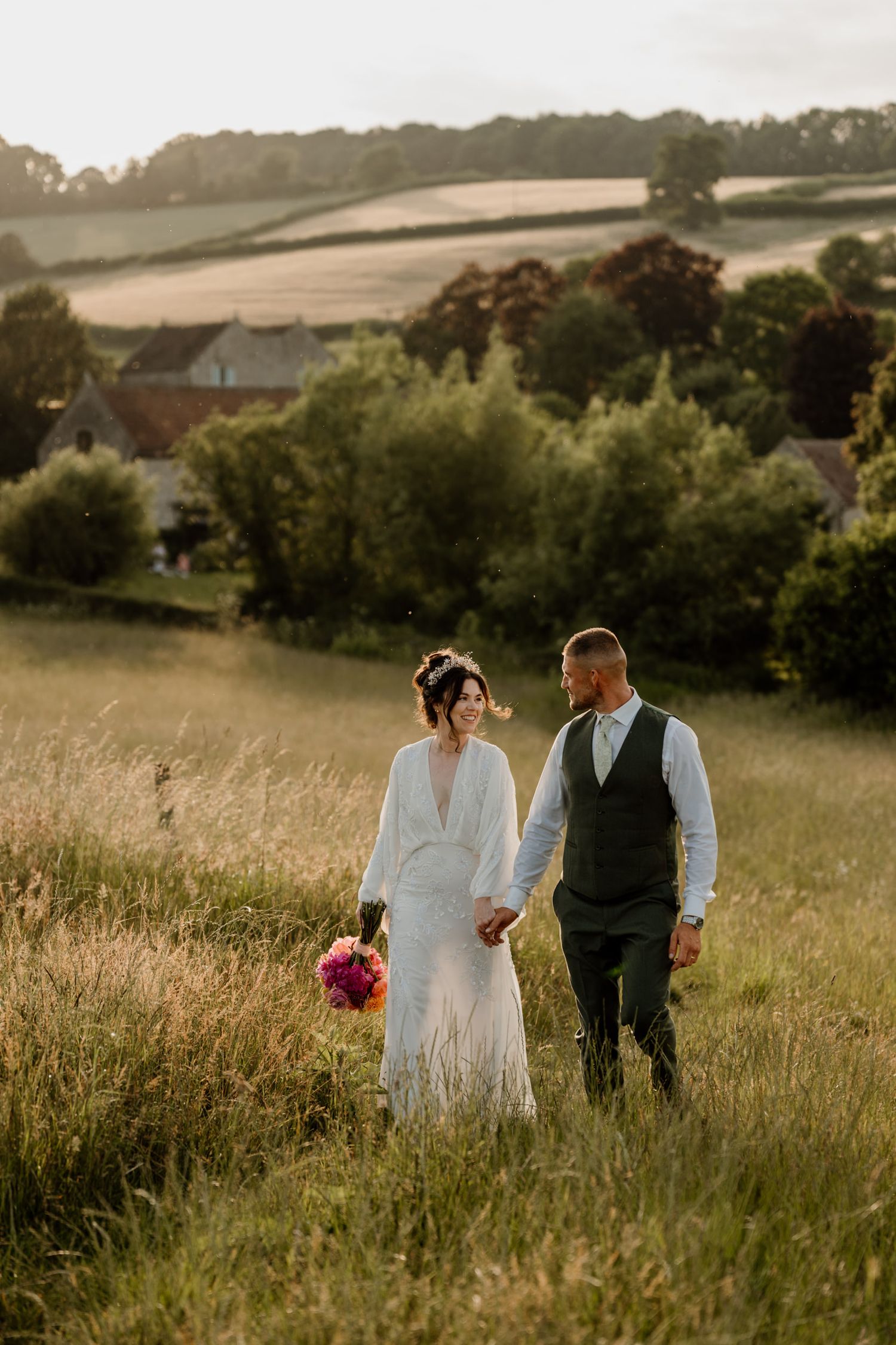Couple walking hand in hand through a rolling meadow during their wedding sunset photos.
