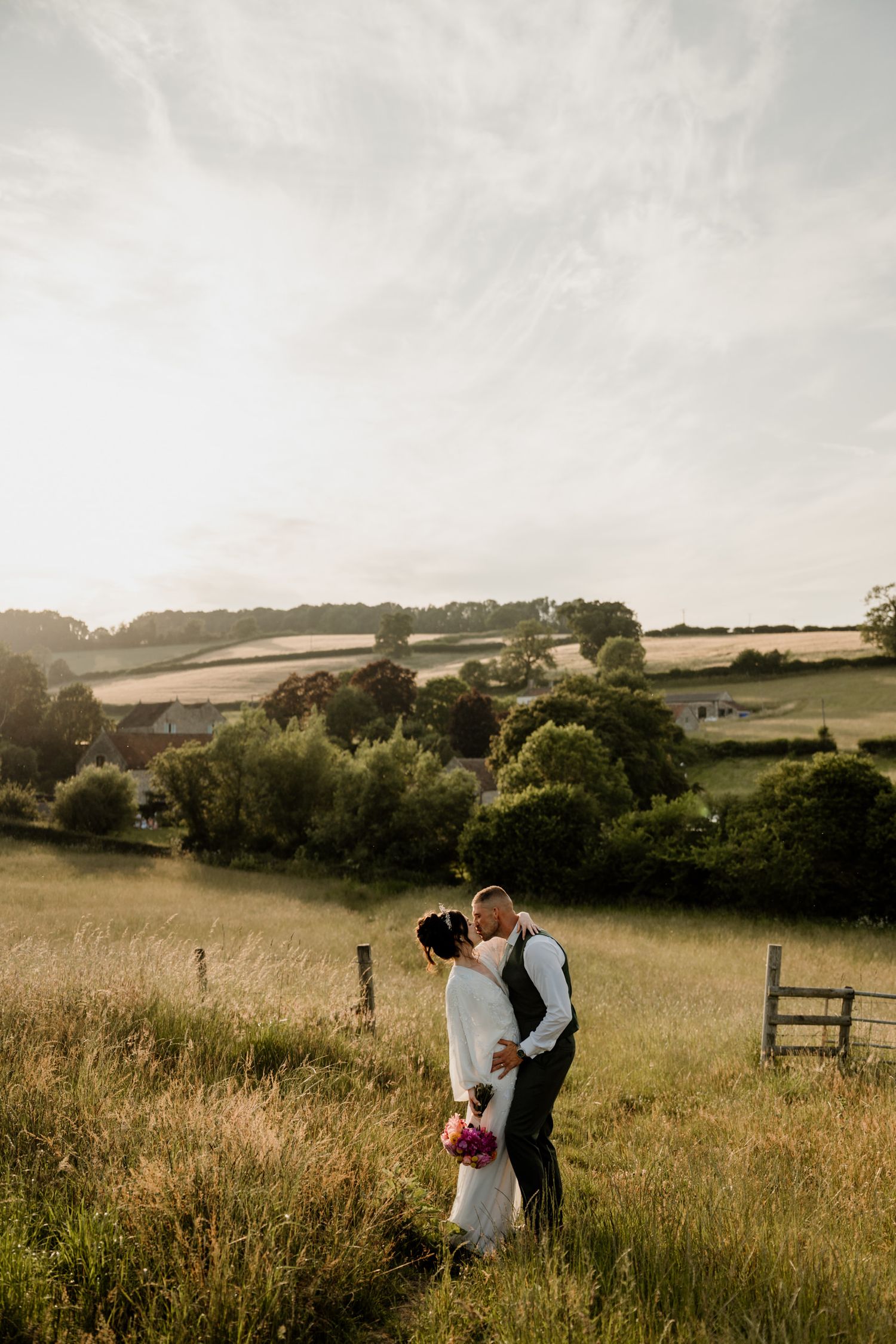 A couple shares an intimate moment in a golden meadow at sunset with rolling hills in the background at Priston Mill in Bath.